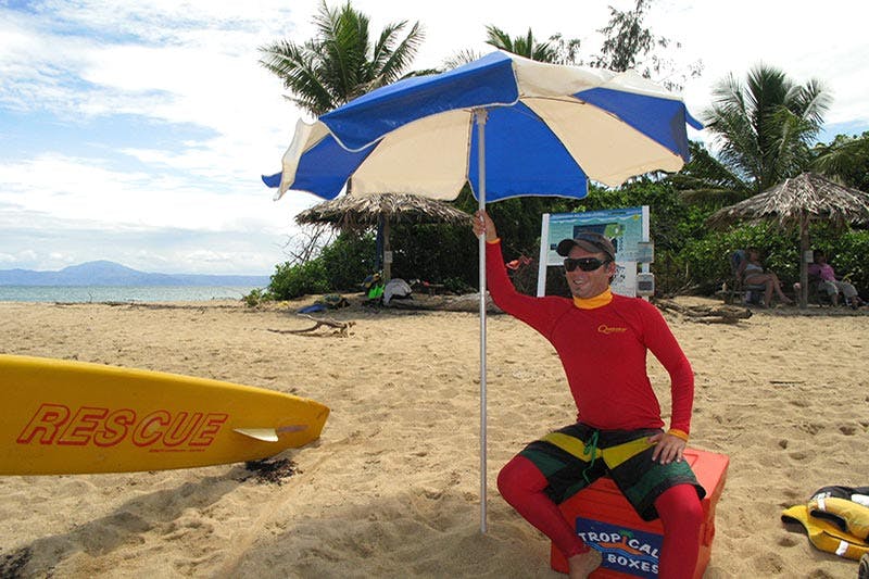 A person in a red rash guard sits under a blue and white umbrella on a sandy beach, with palm trees and people in the background.