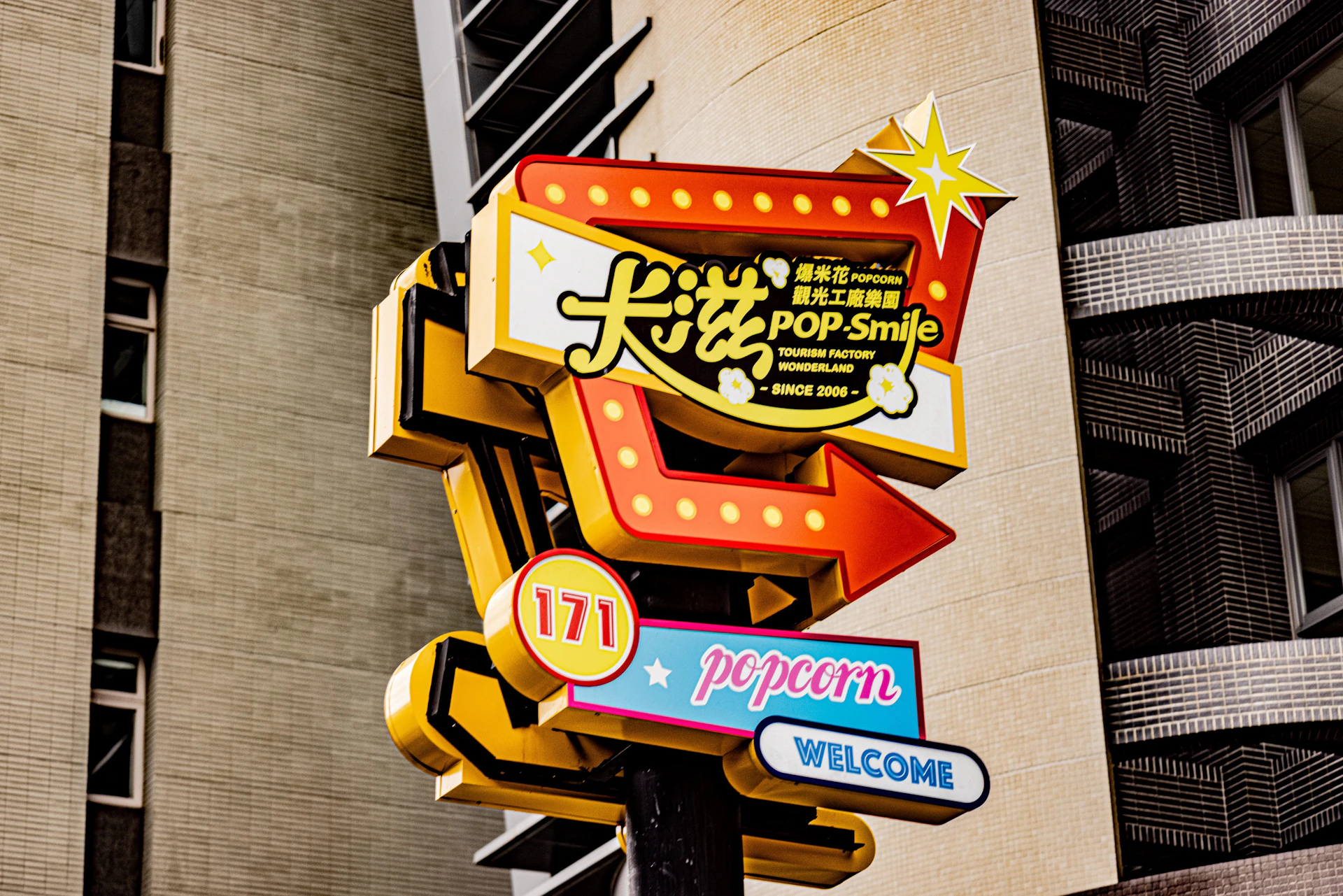 Colorful popcorn-themed sign reading 'POP Smile' with decorative lettering and lights, mounted on a pole in an urban setting.