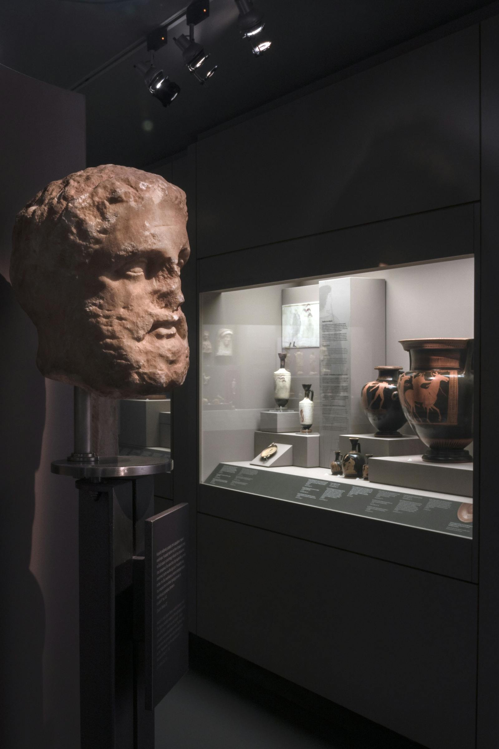 A museum display featuring a sculpted head on a stand and a glass case with ancient pottery and artifacts.