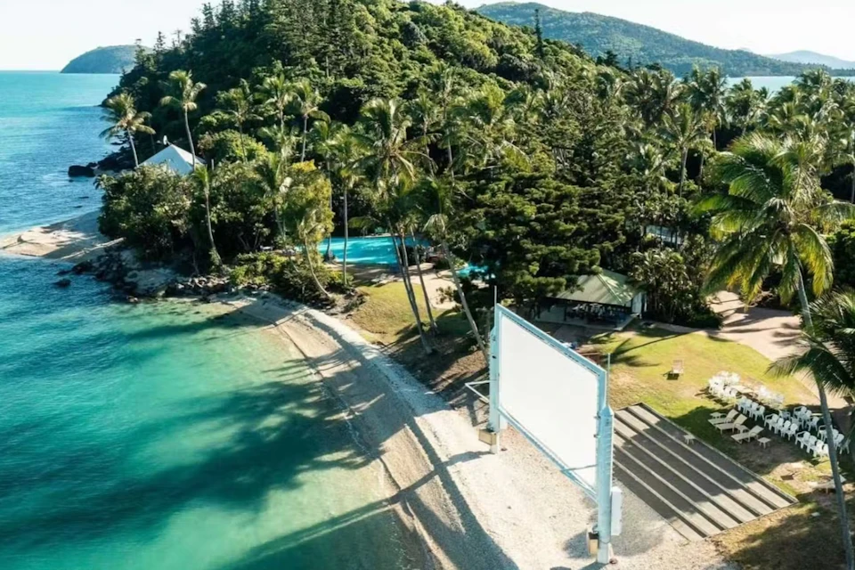 Aerial view of a sandy beach with clear water, surrounded by lush green trees and a two-story building, with a white signboard on the shore.