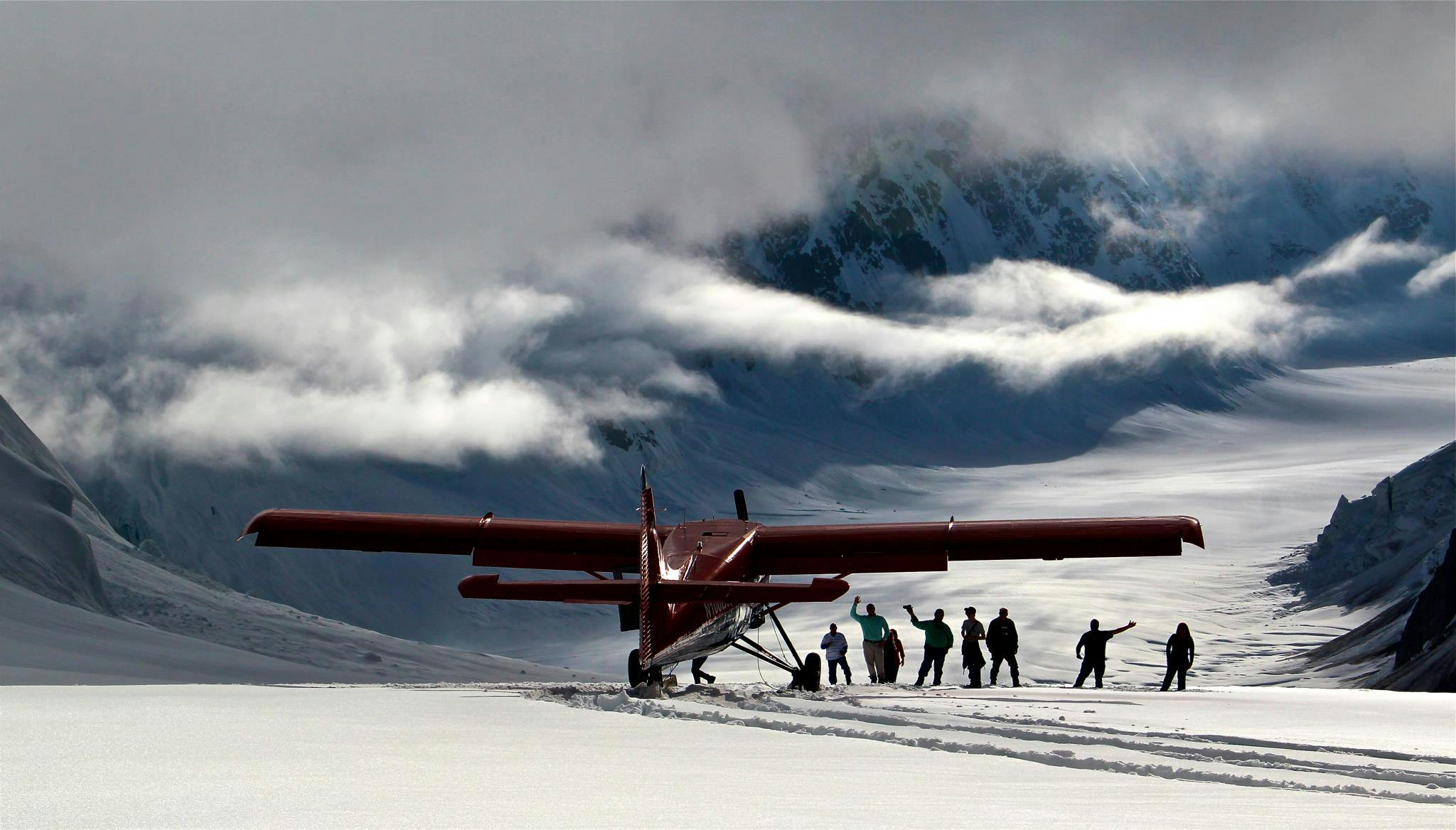 Un petit avion rouge est garé sur un terrain enneigé avec des montagnes en arrière-plan. Plusieurs personnes se tiennent à proximité, certaines faisant un signe de la main.