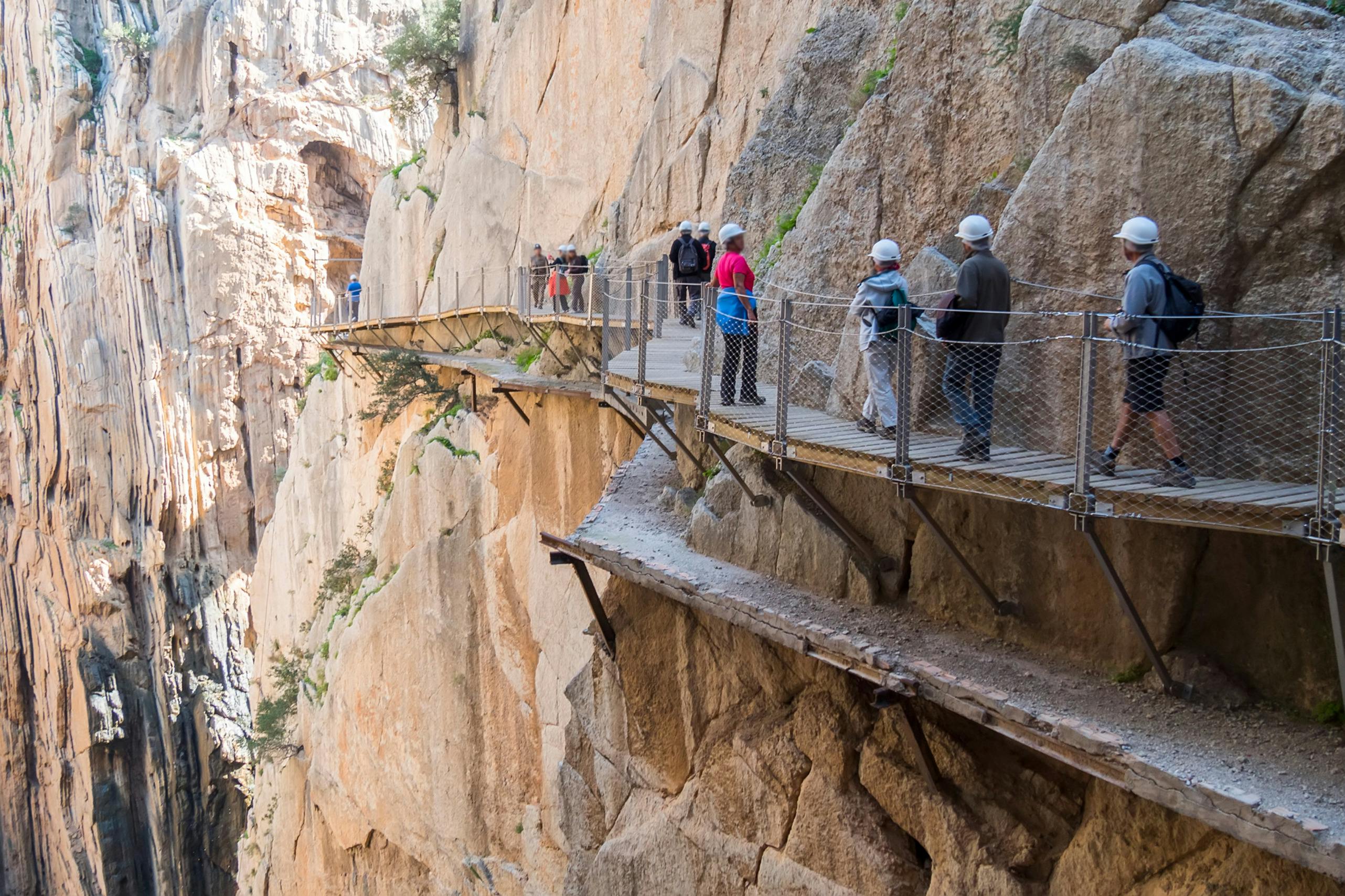 People wearing helmets walk along a narrow, elevated path built into steep cliff walls. A safety rail runs along the path.