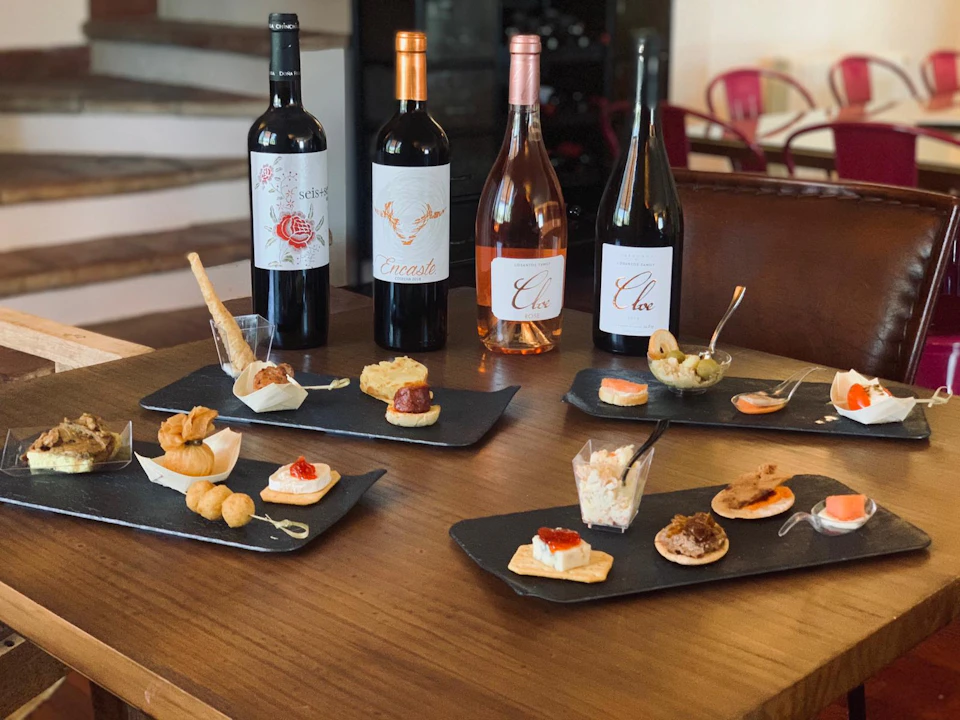 A selection of appetizers on a wooden table with four wine bottles in the background, against a cozy indoor setting.