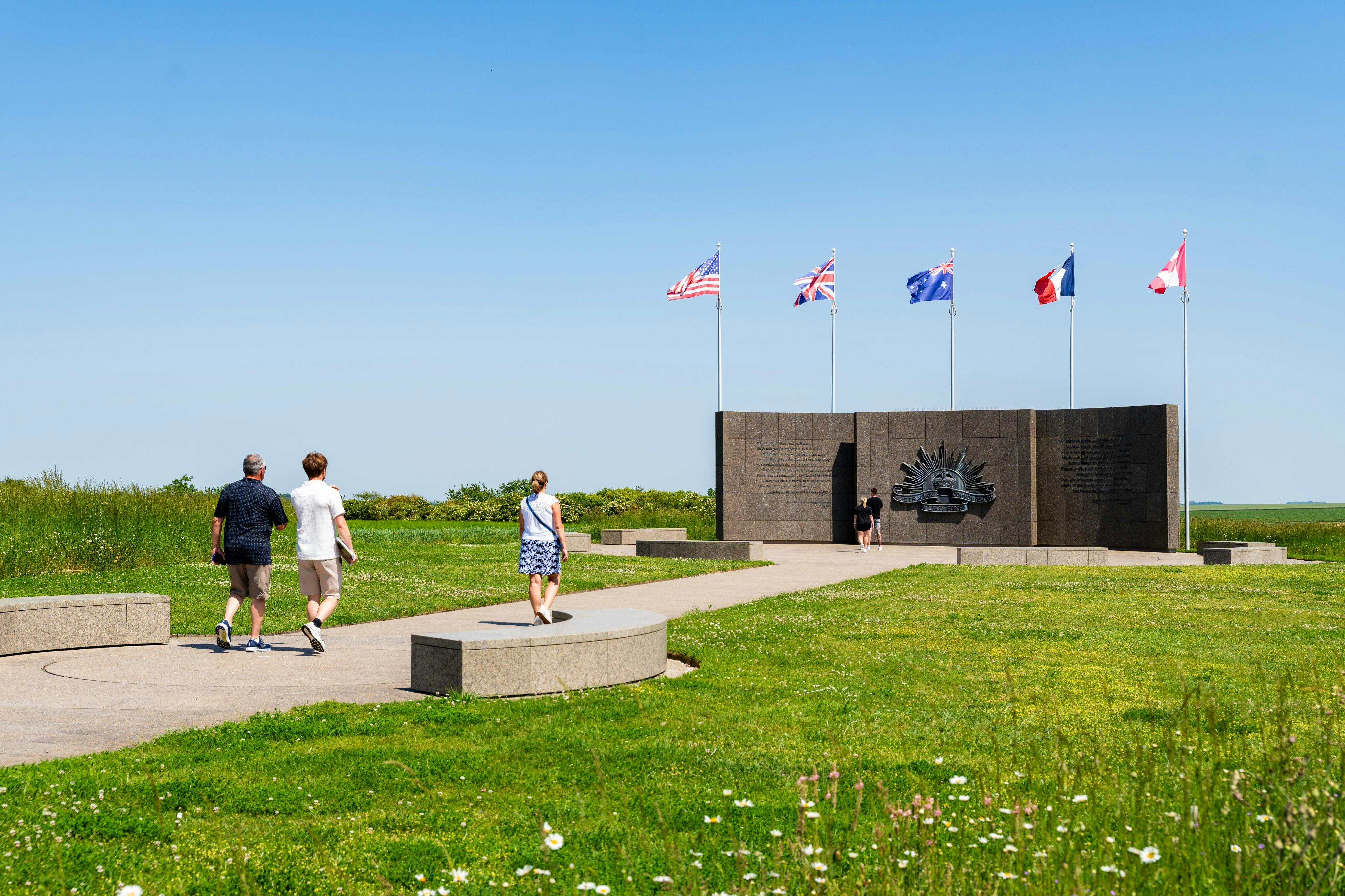 People walking towards a memorial structure with flags of several nations and a well-maintained grassy area under a clear sky.