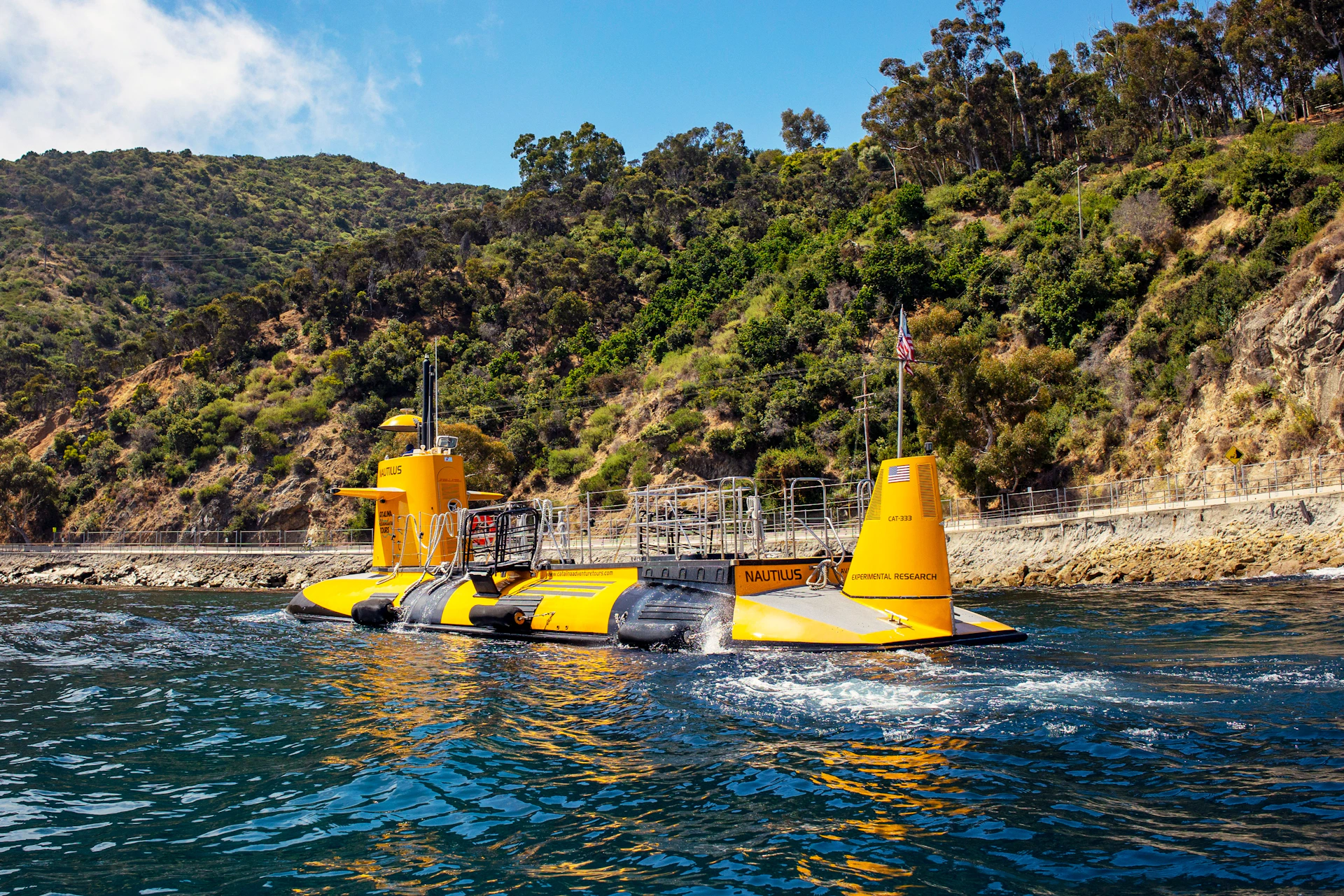 Bright yellow submarine titled "Nautilus" floating near a rocky, tree-covered coast under a blue sky.