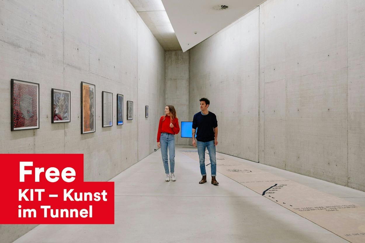 Zwei Personen gehen durch eine Galerie für moderne Kunst mit Betonwänden und betrachten die dort ausgestellten, gerahmten Kunstwerke.