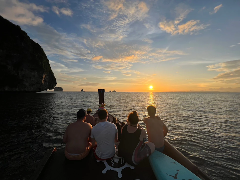 Five people sit on a boat facing the sunset over calm ocean waters with islands visible in the distance.