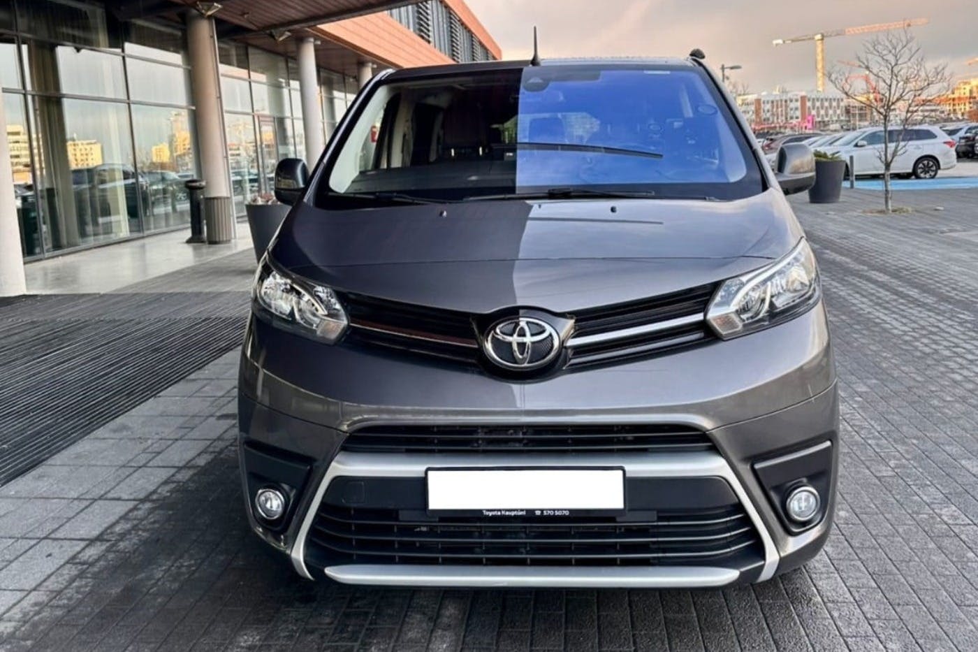 Front view of a parked gray Toyota van in an outdoor urban setting, with buildings and a sidewalk in the background.