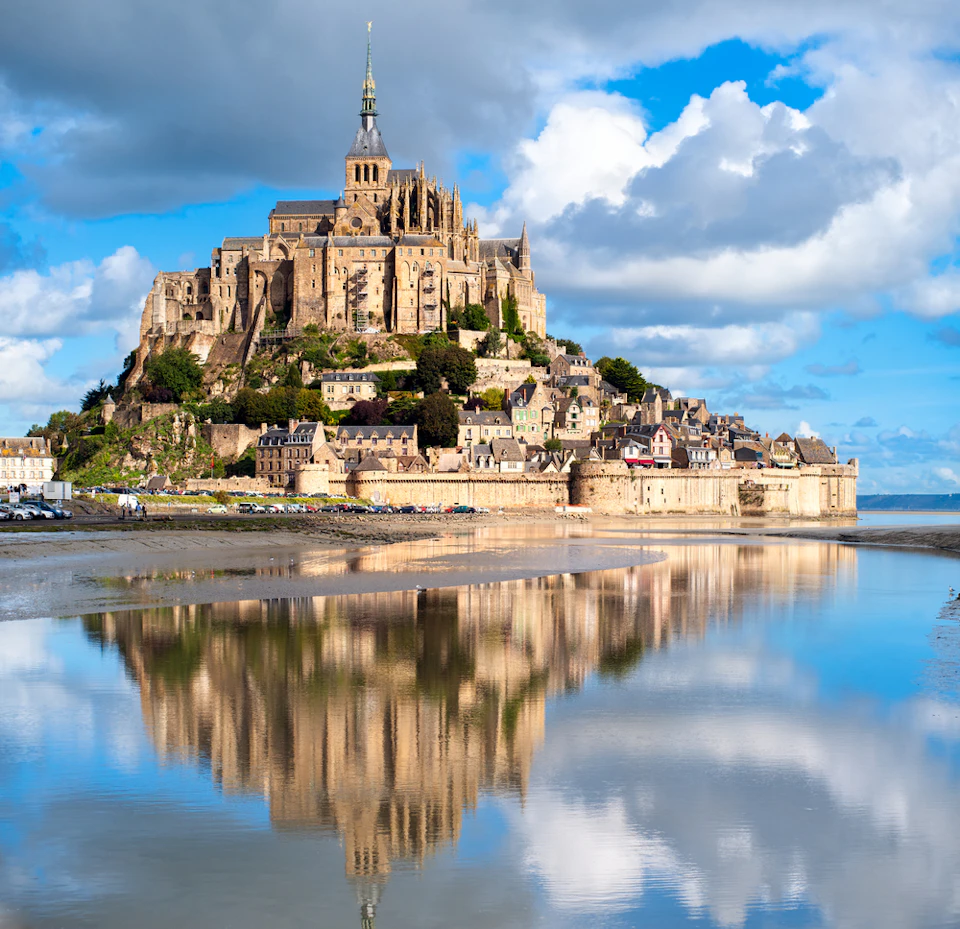 Mont Saint-Michel with its abbey, surrounded by water and reflecting in the stillness, under a partly cloudy blue sky.