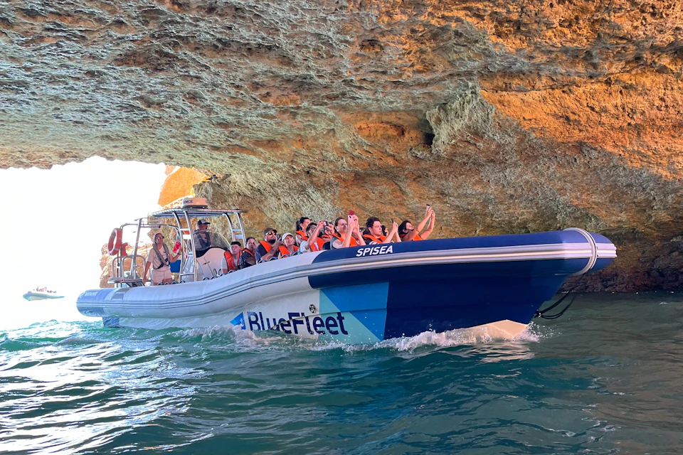 A group of people wearing orange life jackets on a BlueFleet boat, exploring a cave with rocky walls above water.