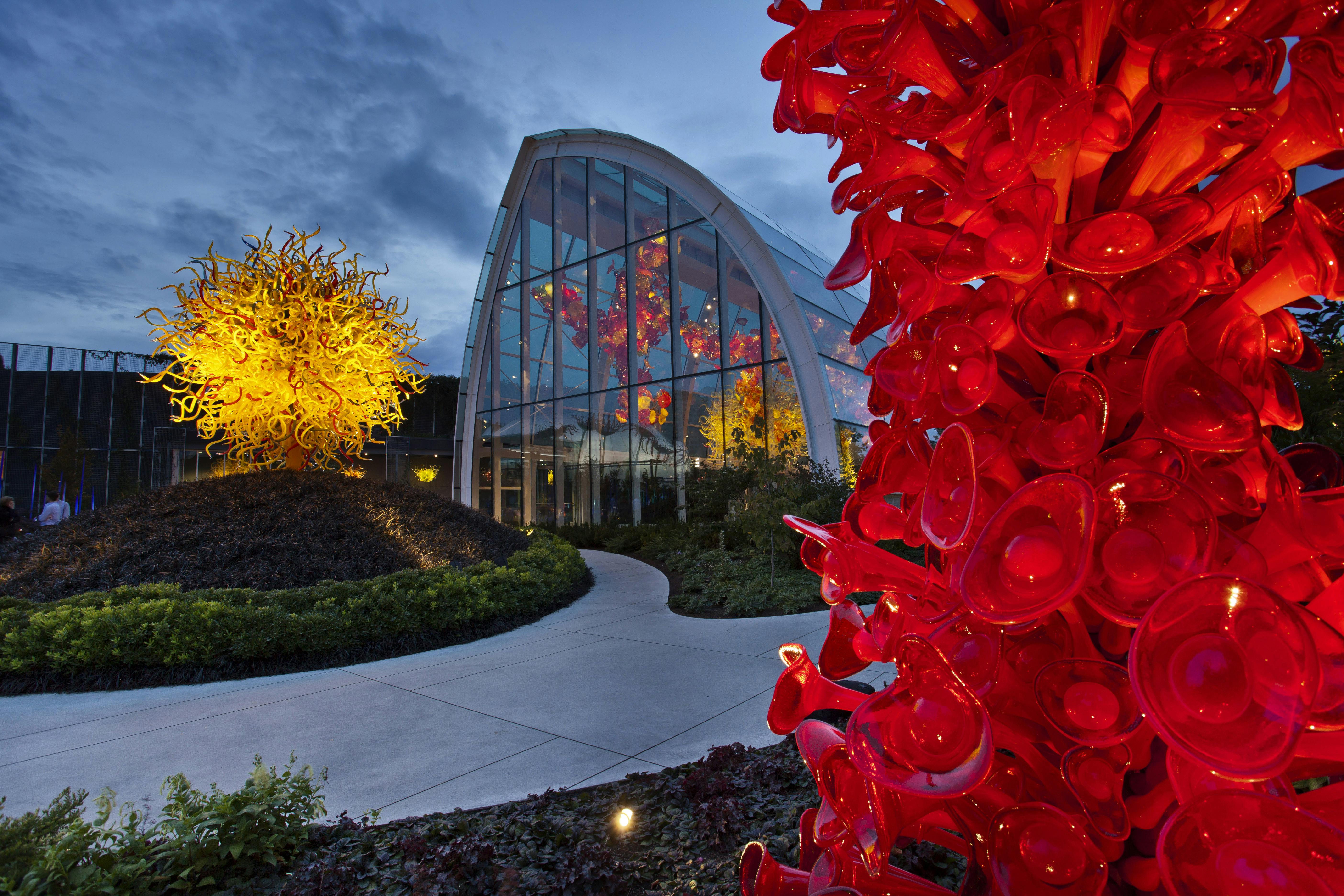玻璃建筑，外面有色彩缤纷的大型玻璃雕塑，在黄昏时点亮。 前景为红色雕塑，背景为黄色。