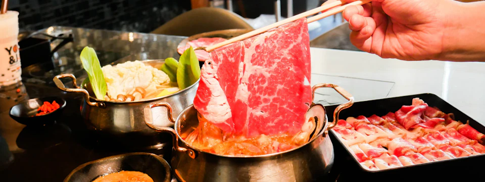 Chopsticks holding a slice of raw, marbled beef over a hot pot with vegetables and broth in a dining setting.