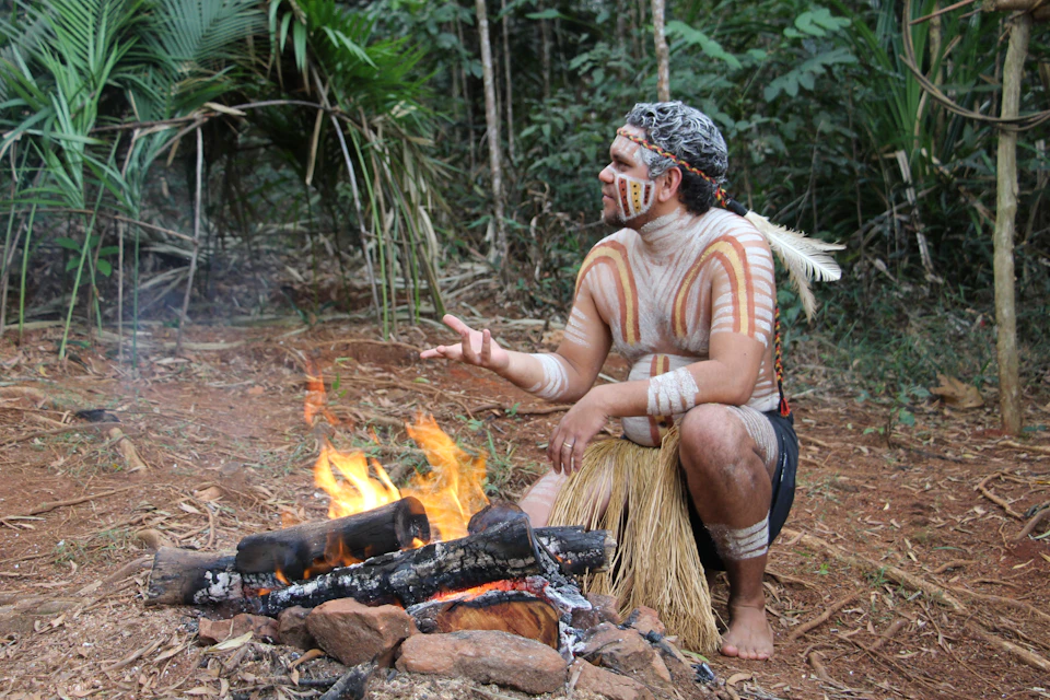 A person with body paint, feathers, and traditional attire sits by a log fire in a forested area, gesturing with their hand.