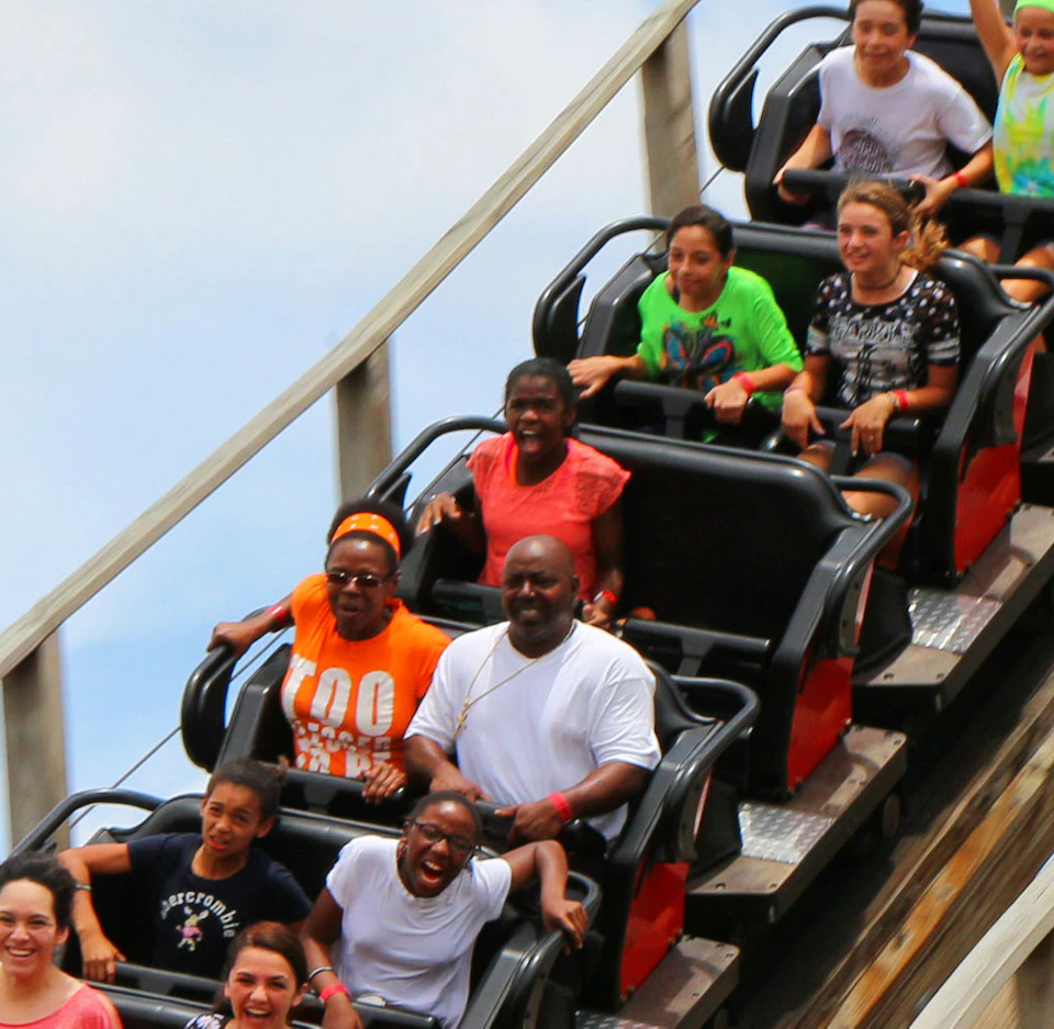 Nine people on a red roller coaster car descending a wooden track, smiling and raising arms against a blue sky.