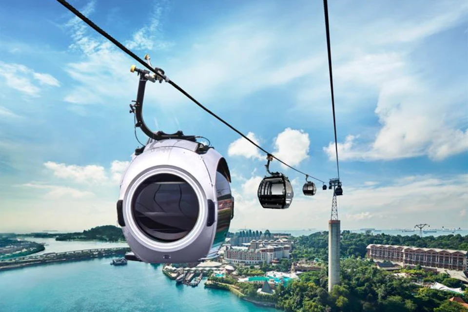 Cable cars with modern designs traveling over water with a cityscape and greenery in the background under a partly cloudy sky.