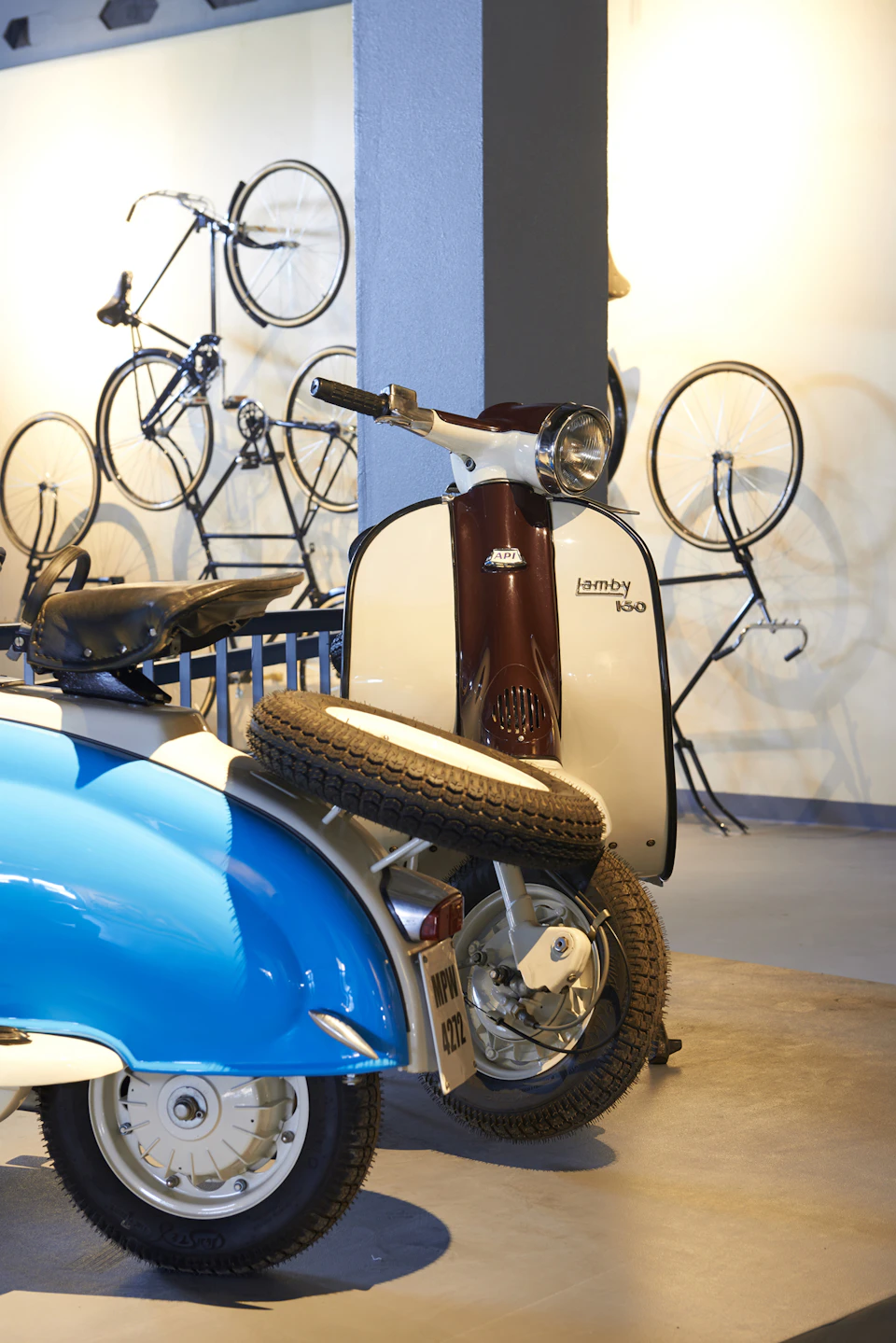 Vintage scooters and bicycles displayed in a well-lit showroom, one scooter in the foreground with a spare tire attached.