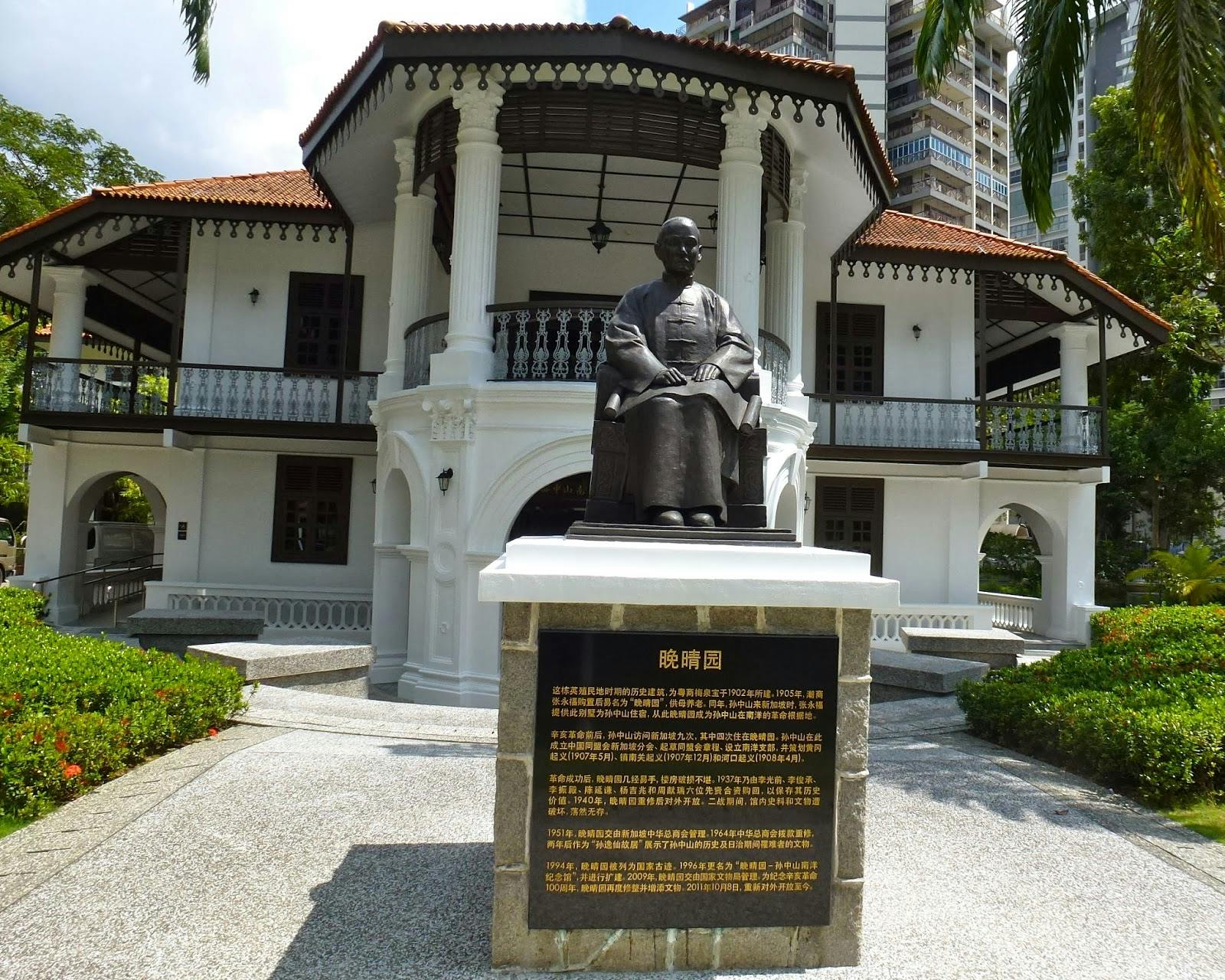 Socha před historickou budovou v bílém koloniálním stylu s cedulí s textem.