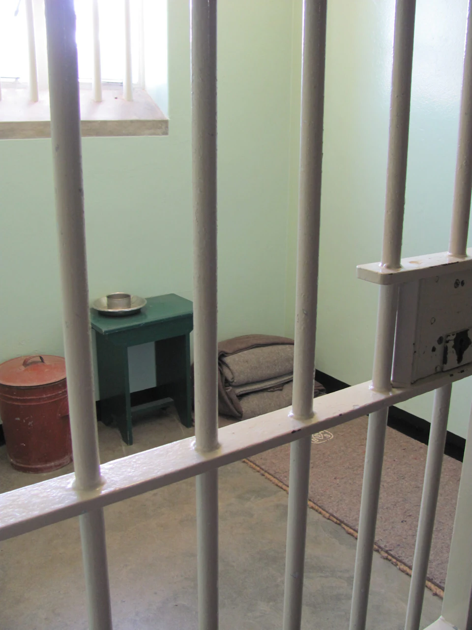A simple prison cell with metal bars, a small table with a metal bowl, a mat on the floor, folded blankets, and a bucket.