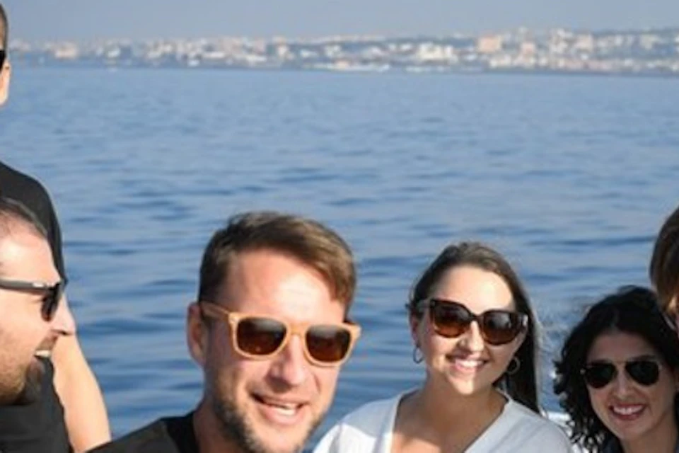 Six people wearing sunglasses, sitting and standing on a boat, smiling with a city skyline in the background.