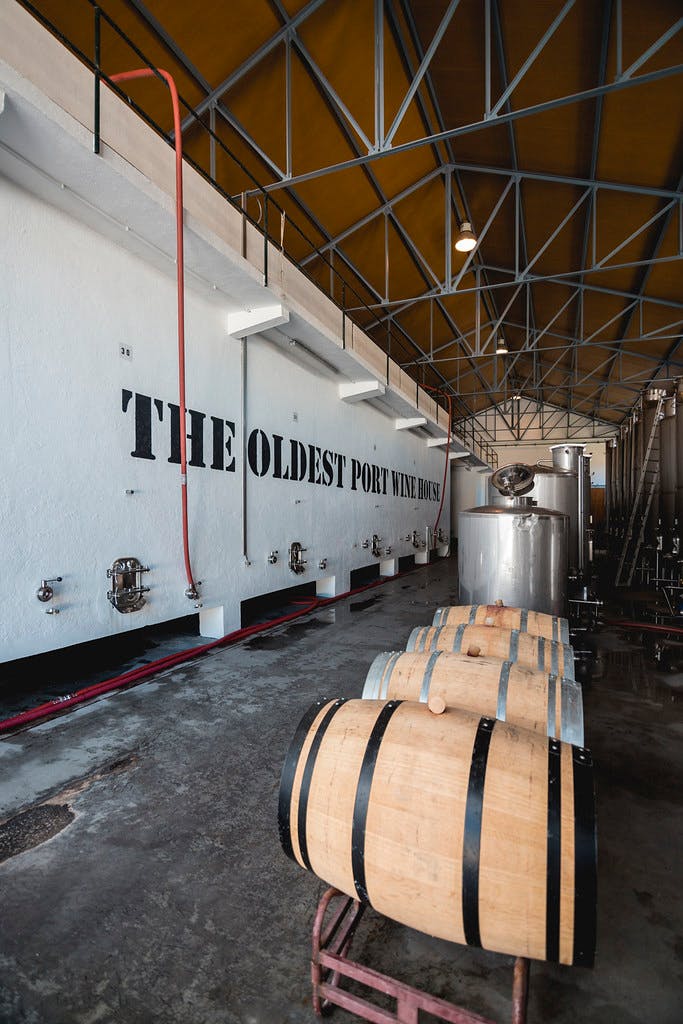 Interieur van een wijnfaciliteit met vaten op de voorgrond en grote tekst op de muur 'The Oldest Port Wine House'.