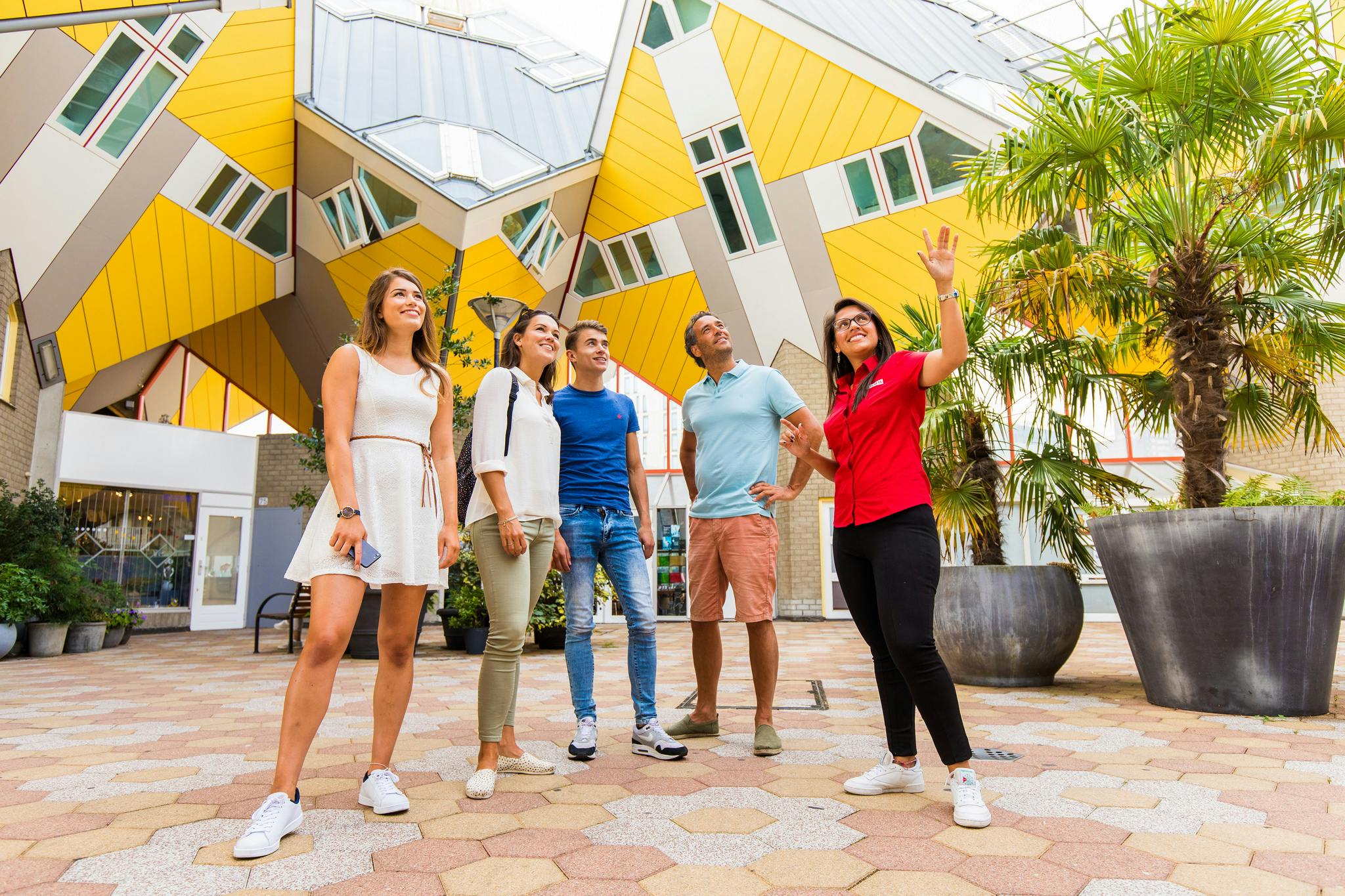 Five people stand smiling and looking upwards, with yellow cube-shaped buildings and potted plants in the background.