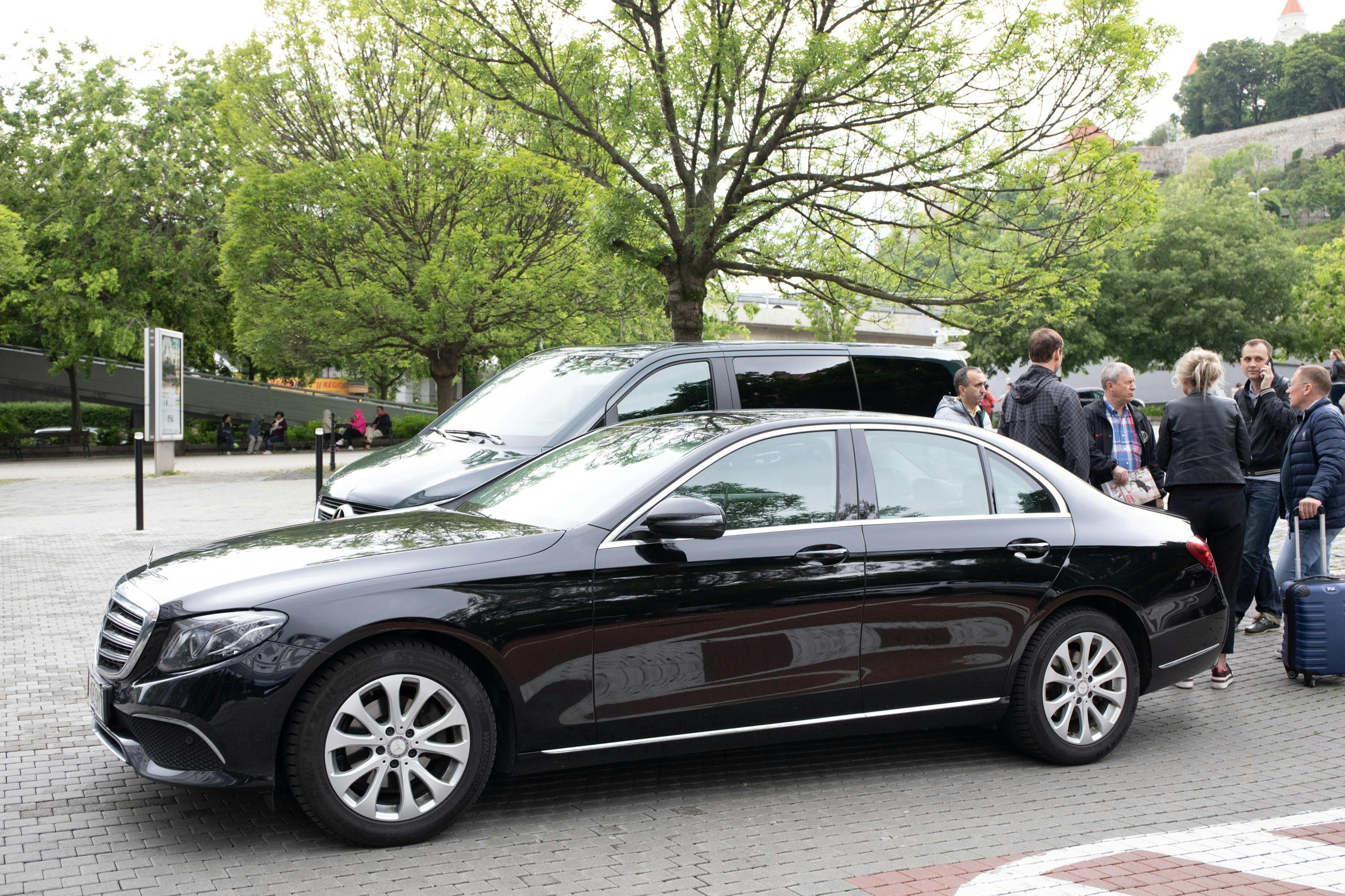 人们站在黑色汽车旁边交谈，停在街道上，后面有树木和山坡。