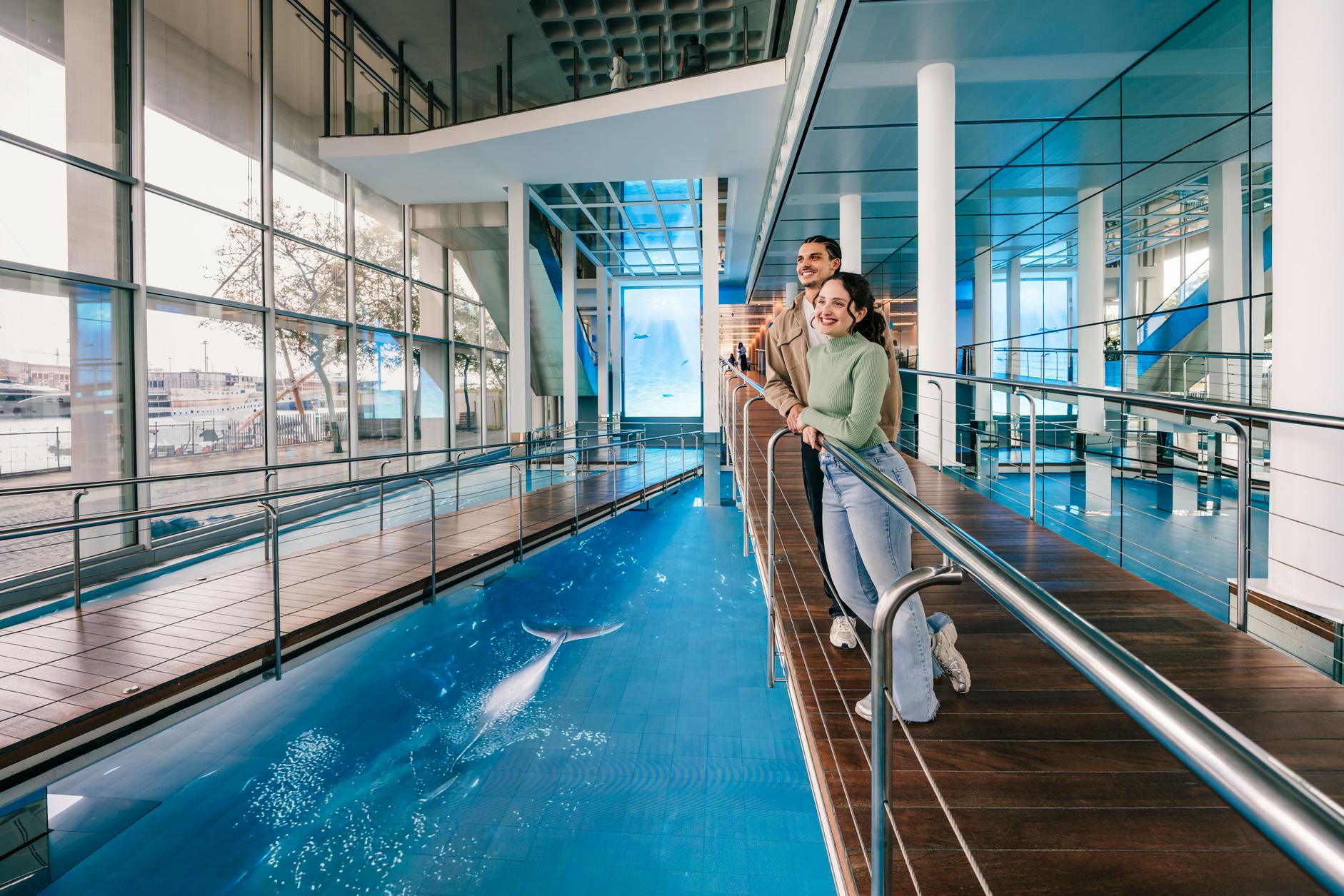 Zwei Personen stehen auf einer hölzernen Brücke in einem modernen Innenraum mit Glaswänden und einem Pool, der eine Wal-Illustration zeigt.