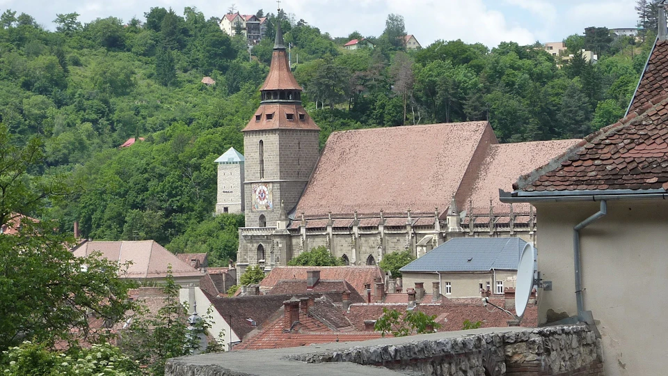 A large stone church with a red-tiled roof and bell tower is surrounded by tree-covered hills and buildings with similar roofs.
