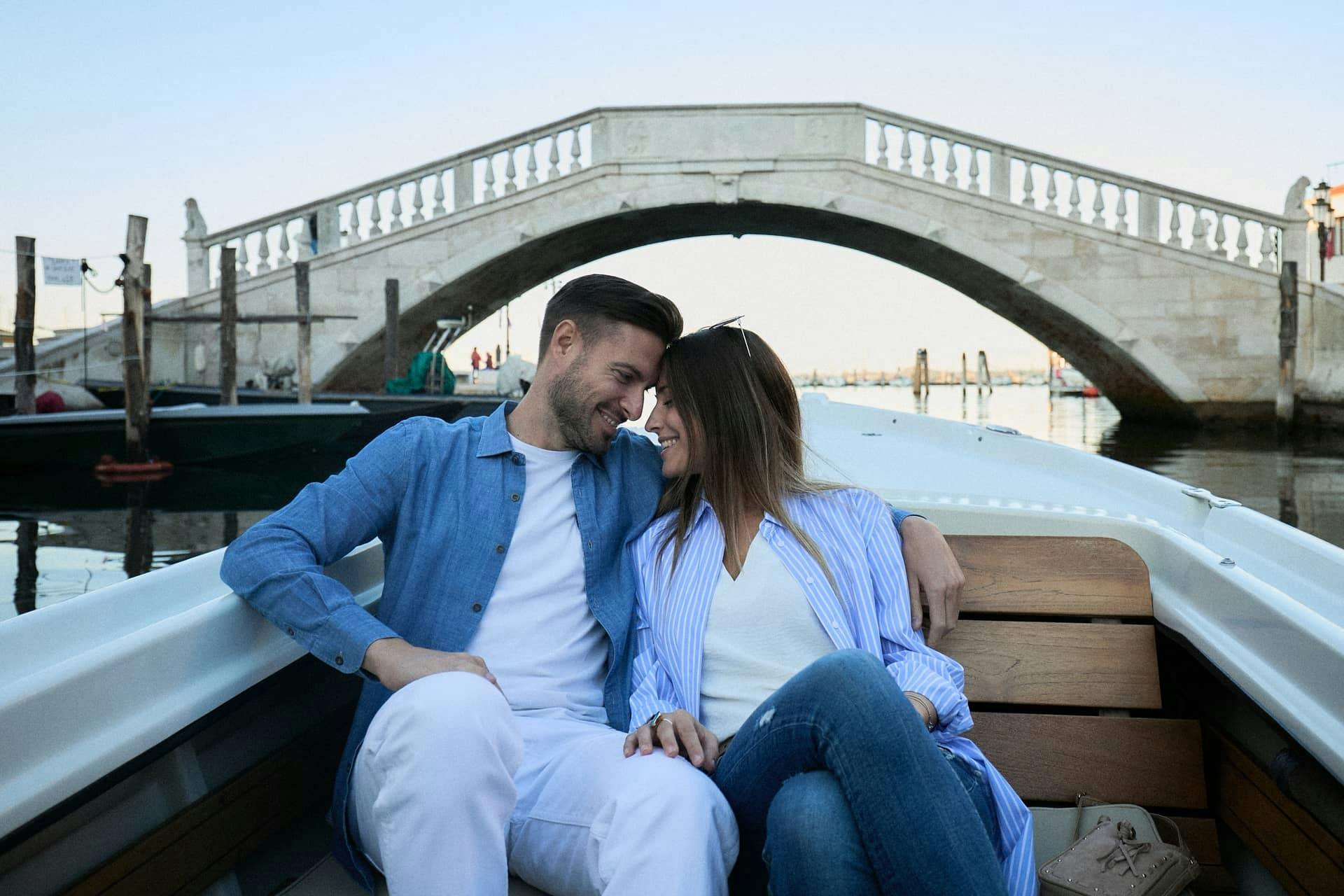 A couple sitting on a boat, smiling at each other, with a stone bridge over a canal in the background.