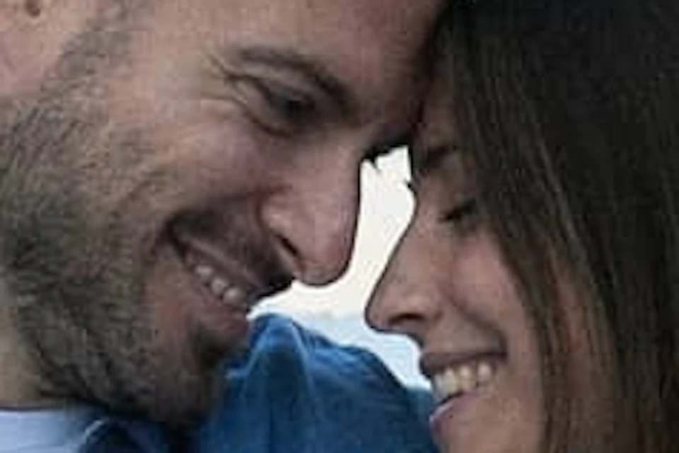 A couple sitting on a boat, smiling at each other, with a stone bridge over a canal in the background.