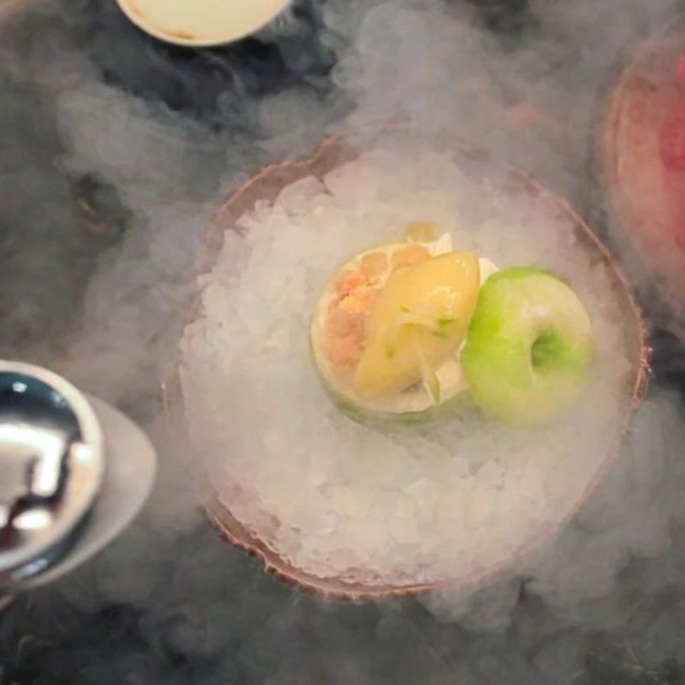 A dish with a green apple and a halved lime, placed on a bed of ice and surrounded by vapor.