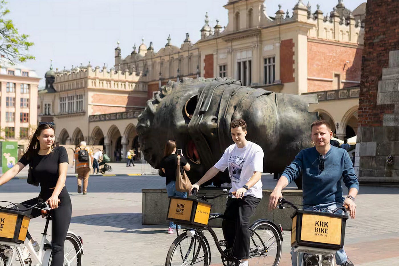 Dva lidé jezdí na kolech v blízkosti velké sochy hlavy na historickém náměstí s ozdobnými budovami v pozadí.