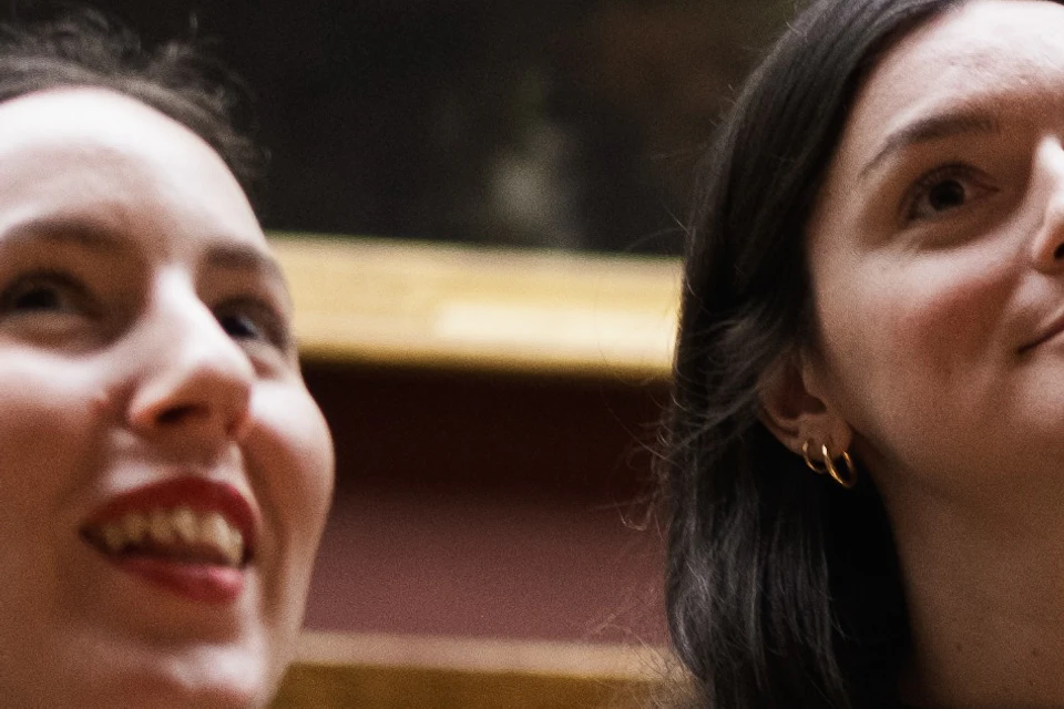 Two women standing in a museum, smiling and looking upward, with framed artwork displayed on the wall behind them.