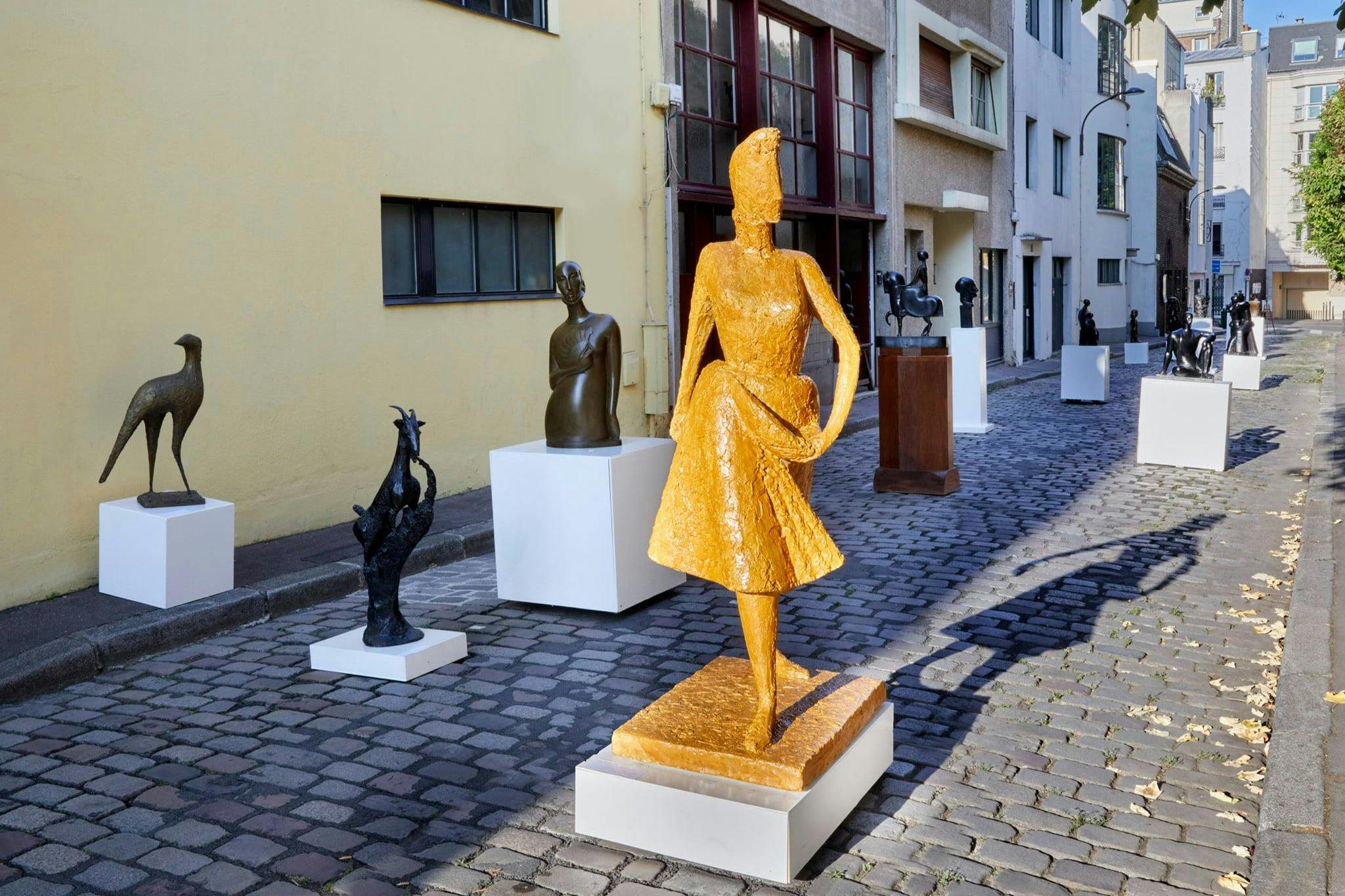 Utomhuskonstutställning av olika skulpturer på piedestaler längs en kullerstensgata intill en gulväggig byggnad.