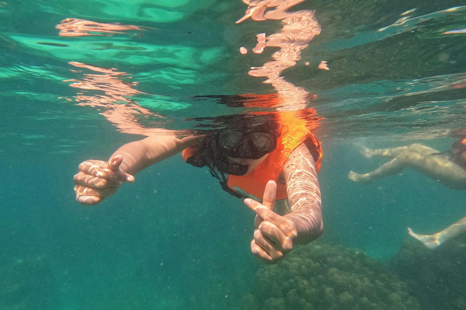 Osoba šnorchlující pod vodou v oranžové záchranné vestě a ukazující oběma rukama palce nahoru.