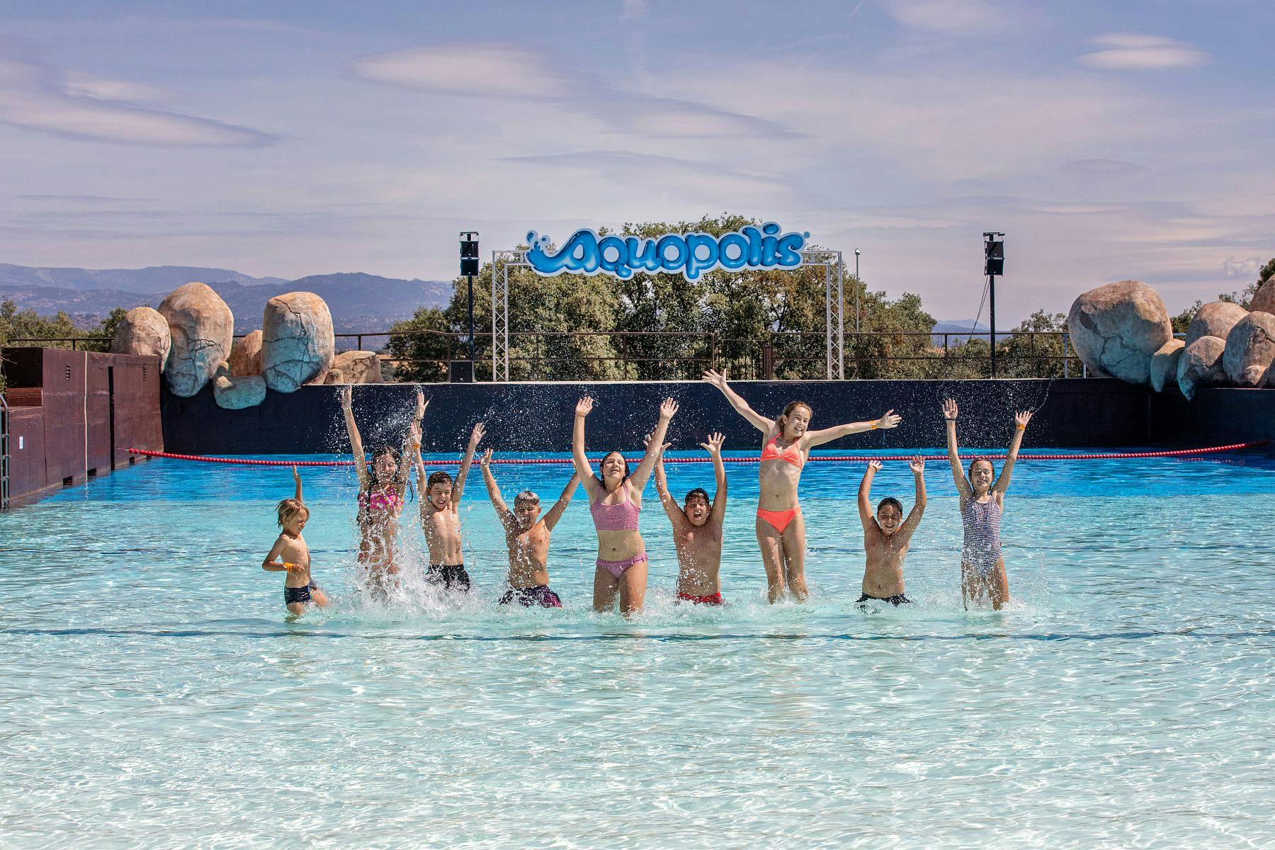 Un grup de persones vestides de bany saltant i esquitxant en una piscina d'un parc aquàtic amb el cartell d'Aquopolis visible al fons.