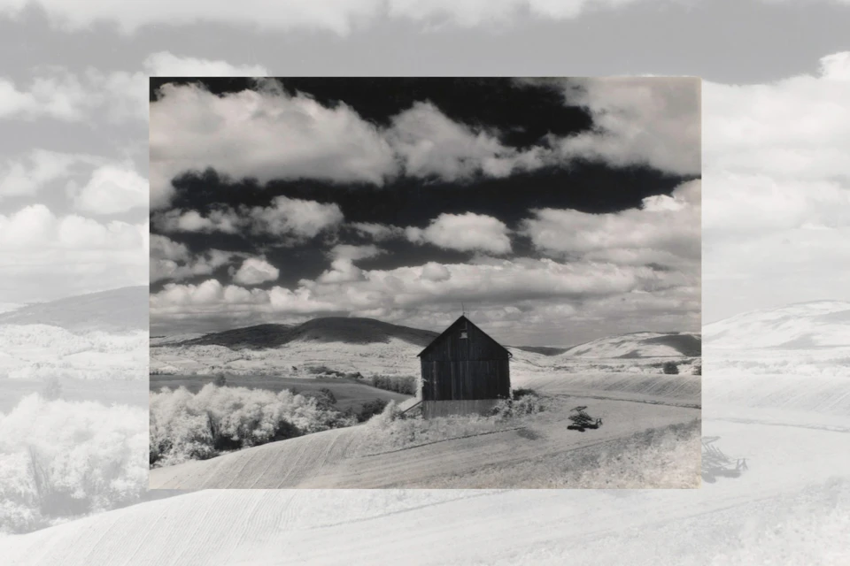 A black-and-white image of a barn on a hill surrounded by fields and rolling hills under a cloudy sky.
