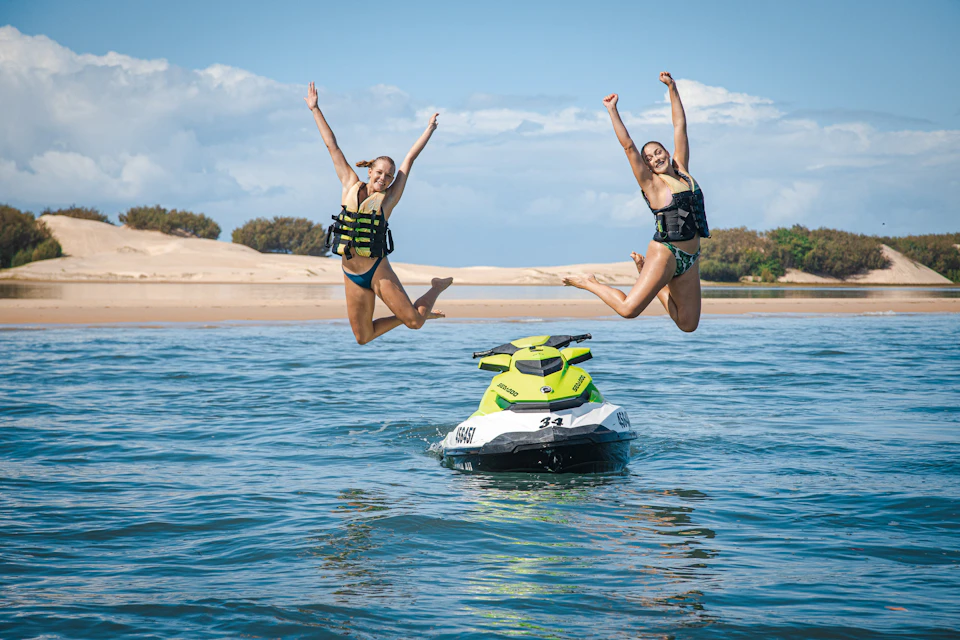 Two women in swimsuits and life vests jump joyfully from a jet ski on clear water with sandy dunes and trees in the background.