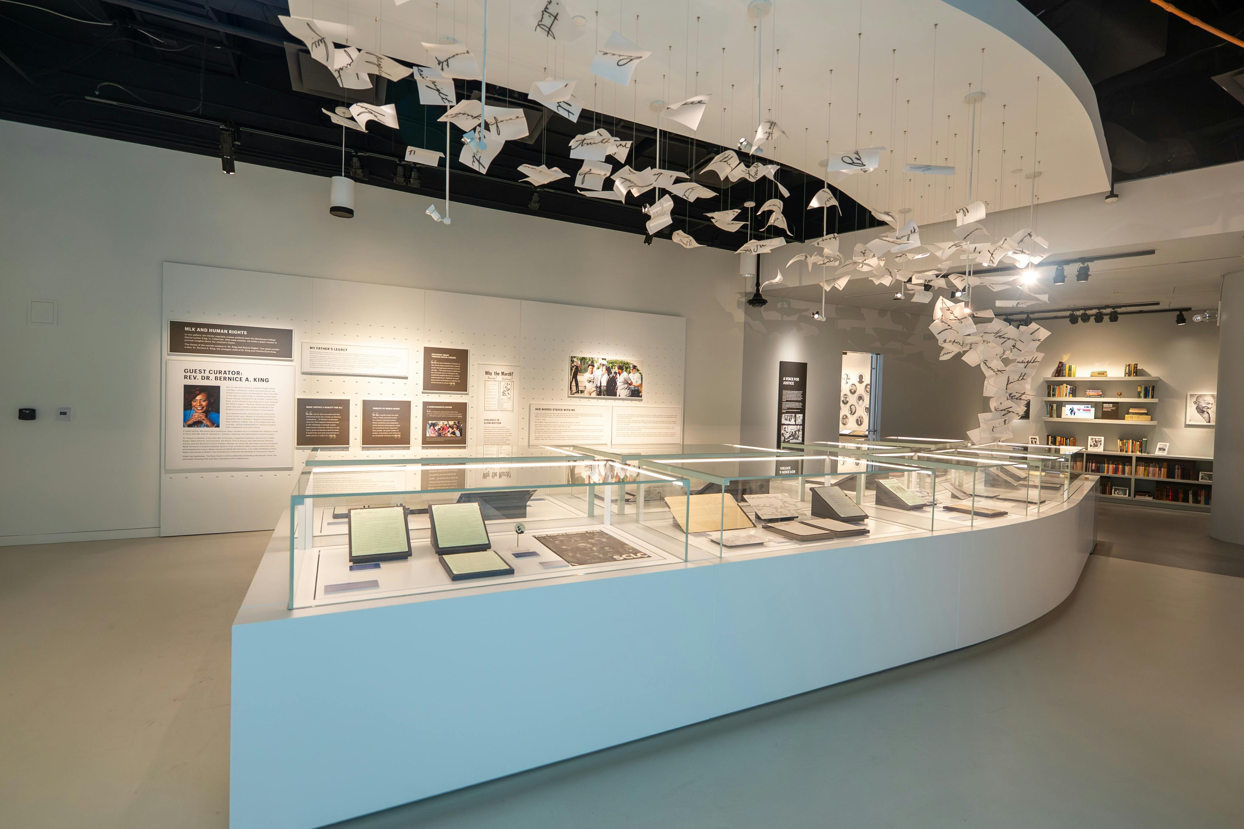 Uma exposição de museu com vitrinas de vidro contendo livros e documentos, e instalações artísticas de papel penduradas por cima.