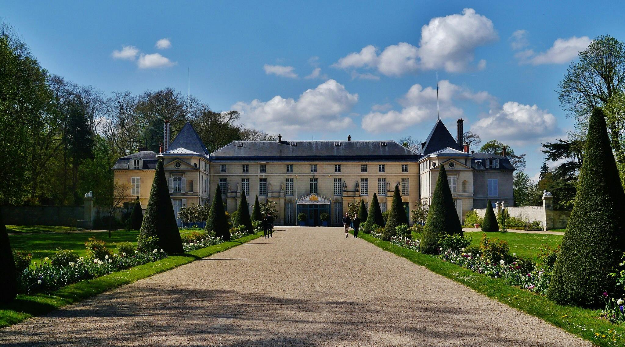 Un grande castello con alberi topiari conici che costeggiano un sentiero di ghiaia, circondato da giardini curati sotto un cielo azzurro e parzialmente nuvoloso.