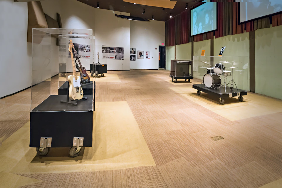 Museum display with electric guitars, a drum kit on wheeled platforms, and vintage music posters on the walls under soft lighting.