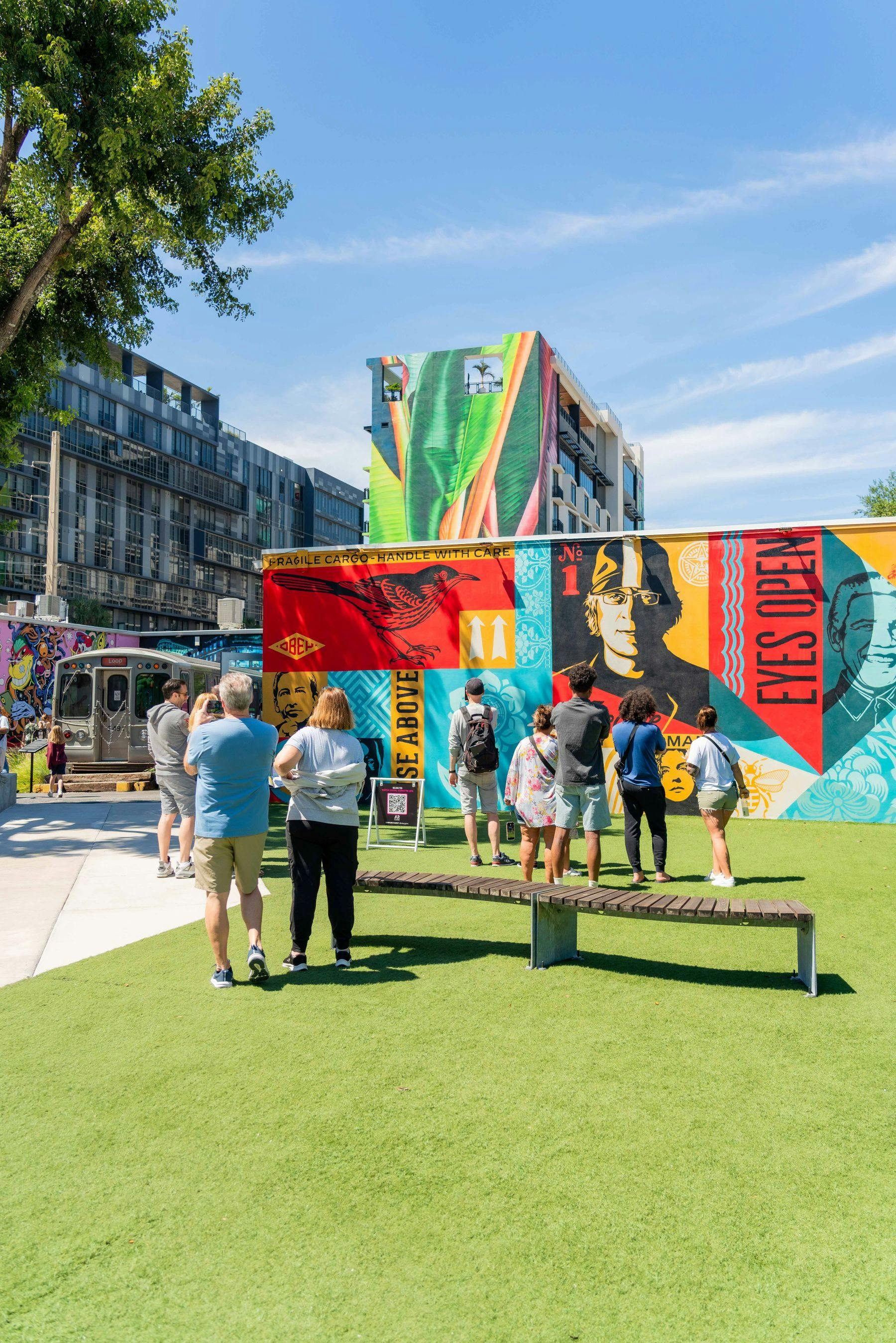 People stand and observe vibrant murals on a building and wall in an outdoor urban area with green artificial grass.