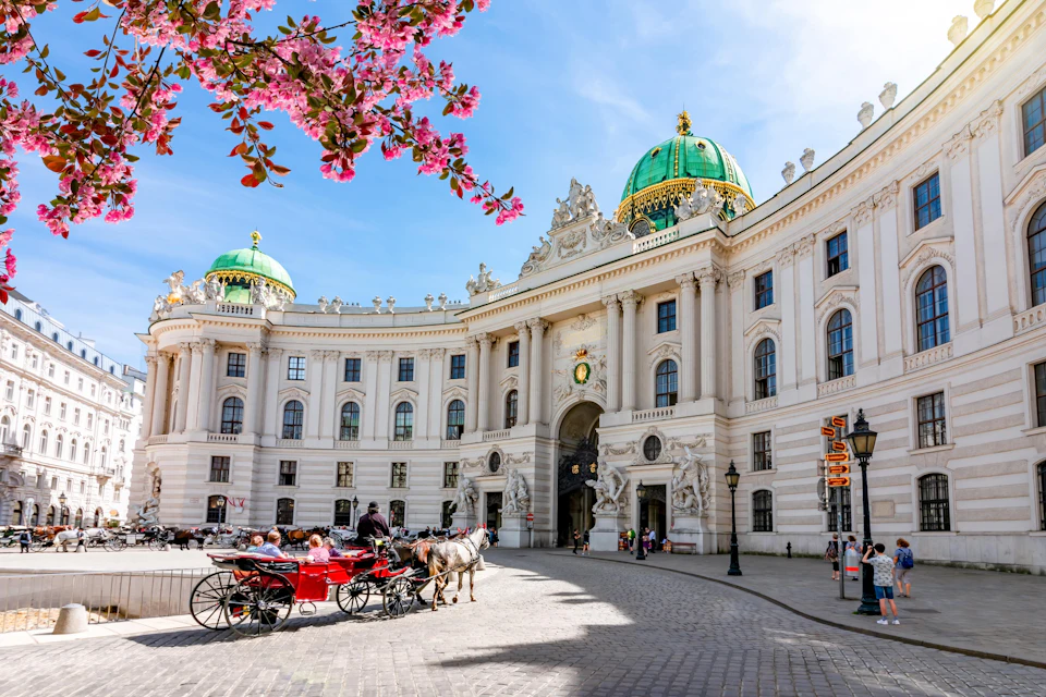 A historic building with a green dome, a horse-drawn carriage, and pink blossoms in the foreground on a sunny day.