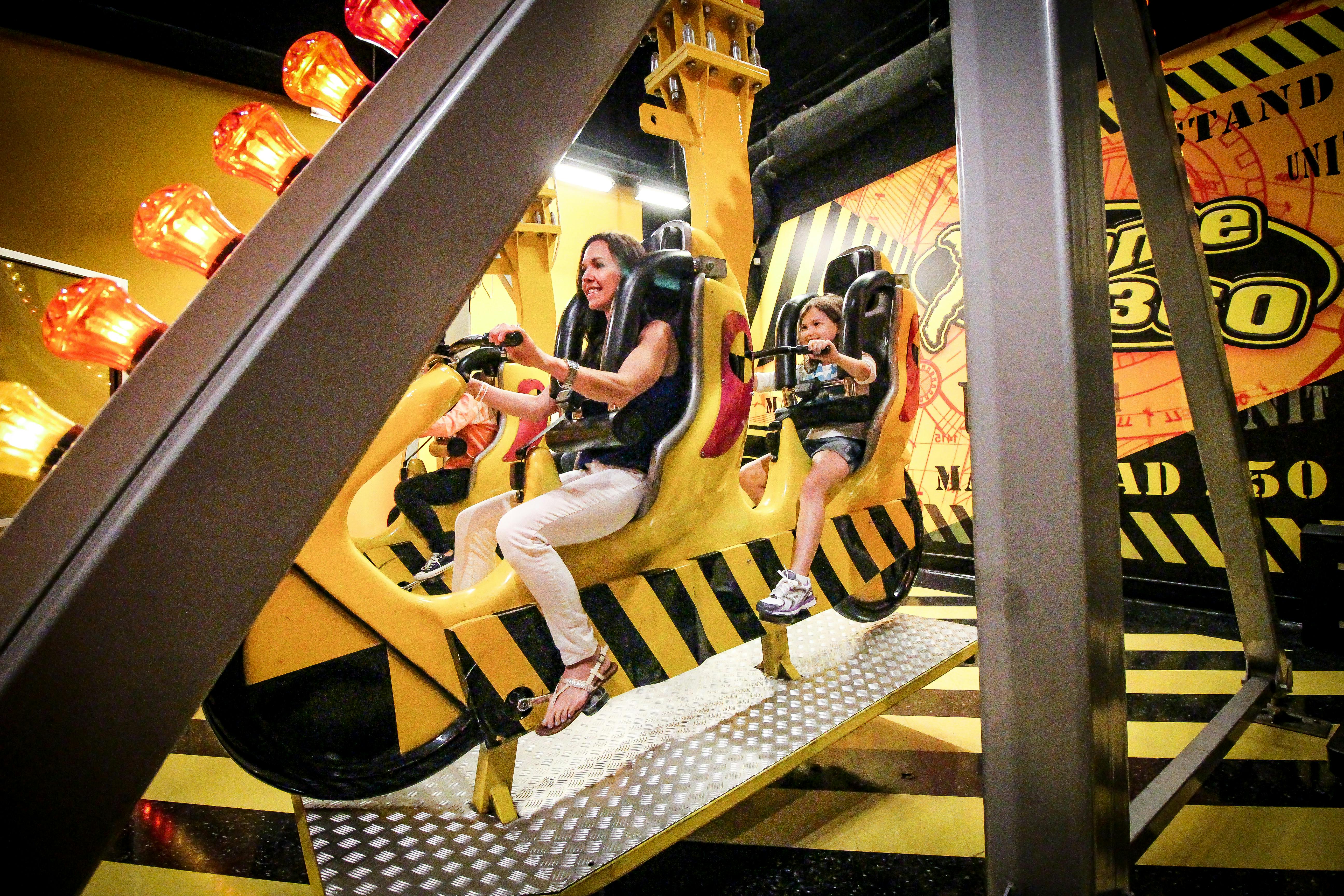 Dva lidé, dospělý a dítě, sedí ve žlutých zábavních sedadlech s bezpečnostními postroji, obklopených průmyslovým dekorem.