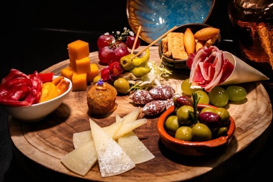 A wooden platter with assorted cheeses, olives, cured meats, crackers, grapes, and skewered appetizers, arranged decoratively.