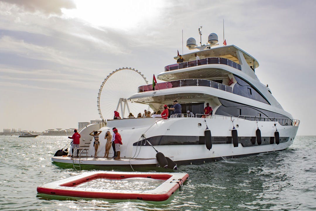 Lidé v plavkách na vícepodlažní jachtě s nedalekým vodním bazénem; v pozadí je vidět ruské kolo.