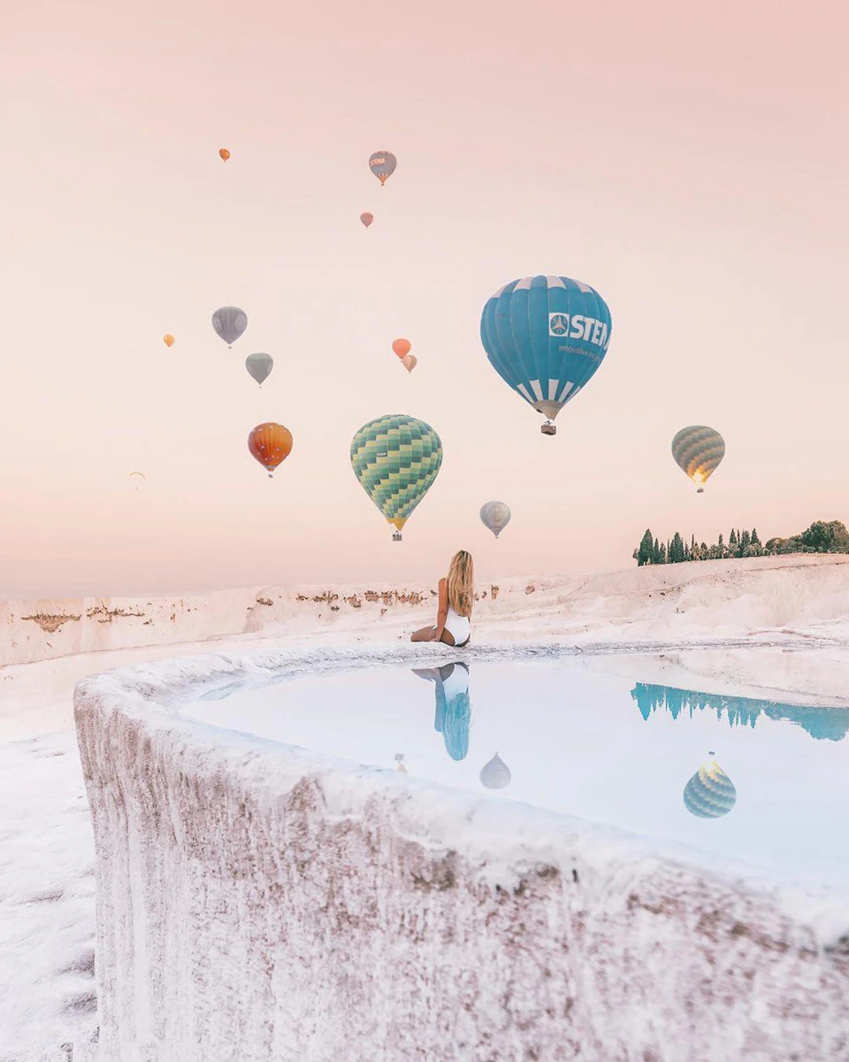 A person in swimwear sits by a reflective pool, watching colorful hot air balloons float in a pastel sky.