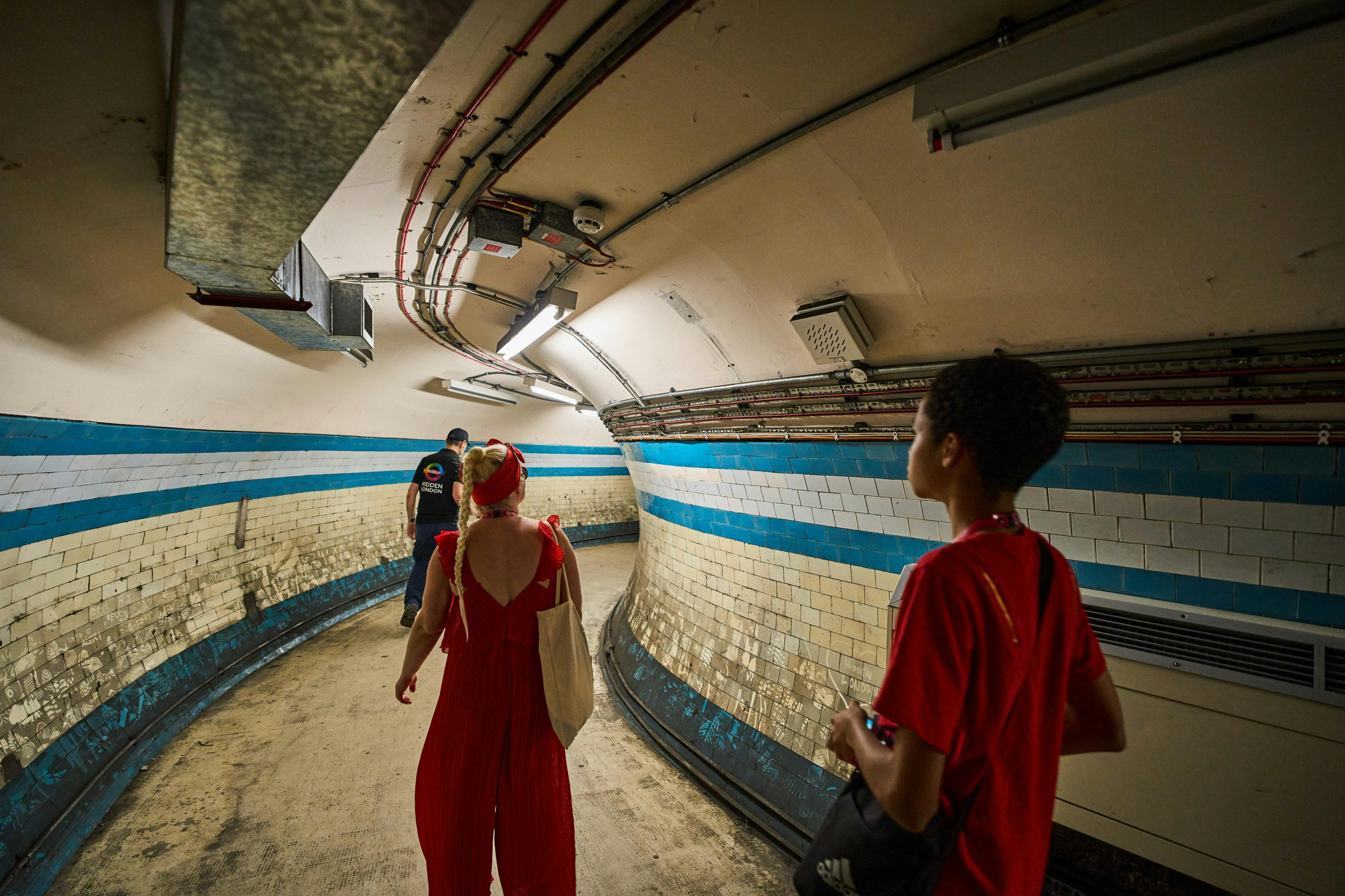 People walking through a curved underground tunnel with tiled walls and overhead lighting.