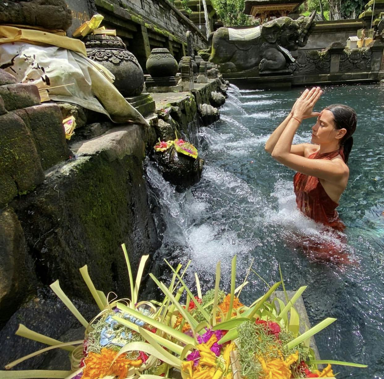 A woman in a red dress prays in a temple pool, surrounded by offerings and stone carvings, with water flowing from spouts.