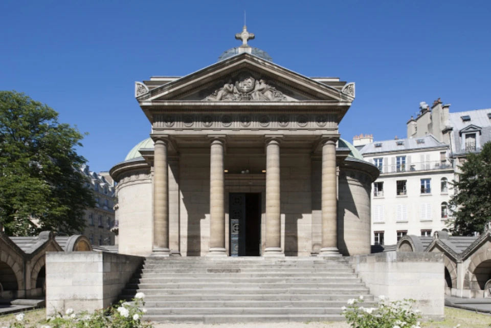A neoclassical-style building with columns, a triangular pediment, and a dome, set against a clear blue sky.